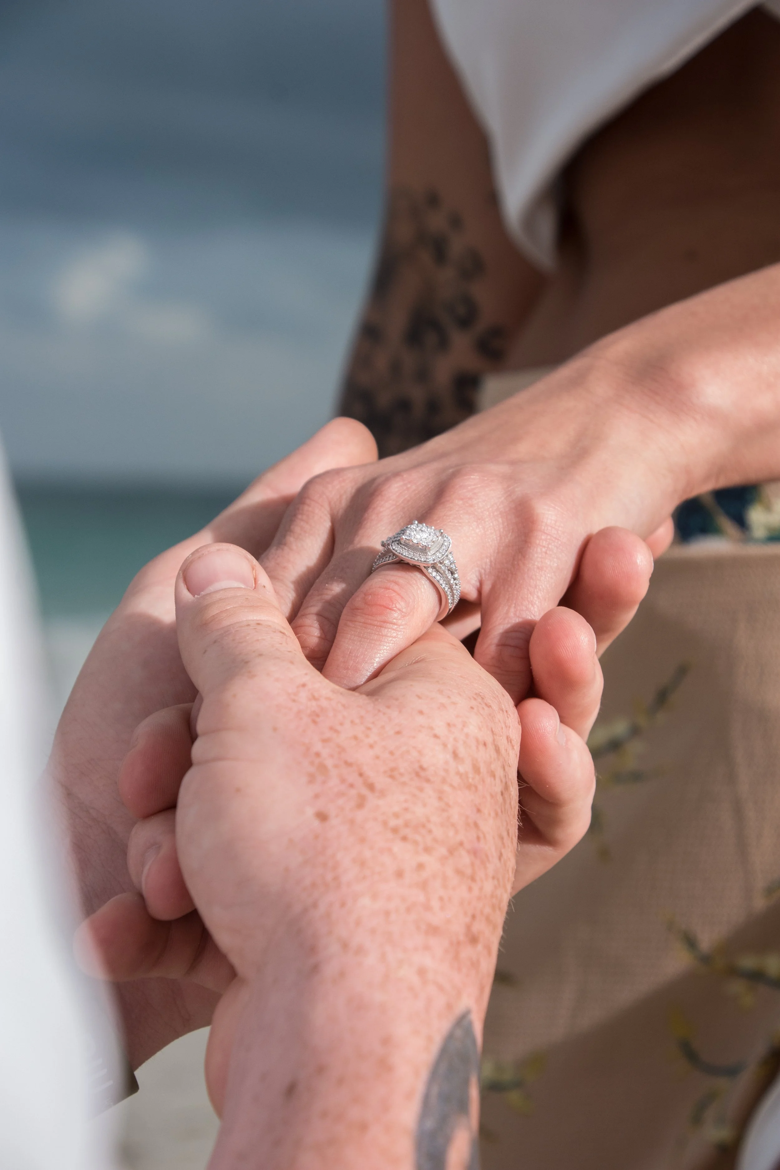 Close-up of a person wearing a diamond engagement ring and wedding band, holding hands with another person at the beach with ocean and cloudy sky in the background.