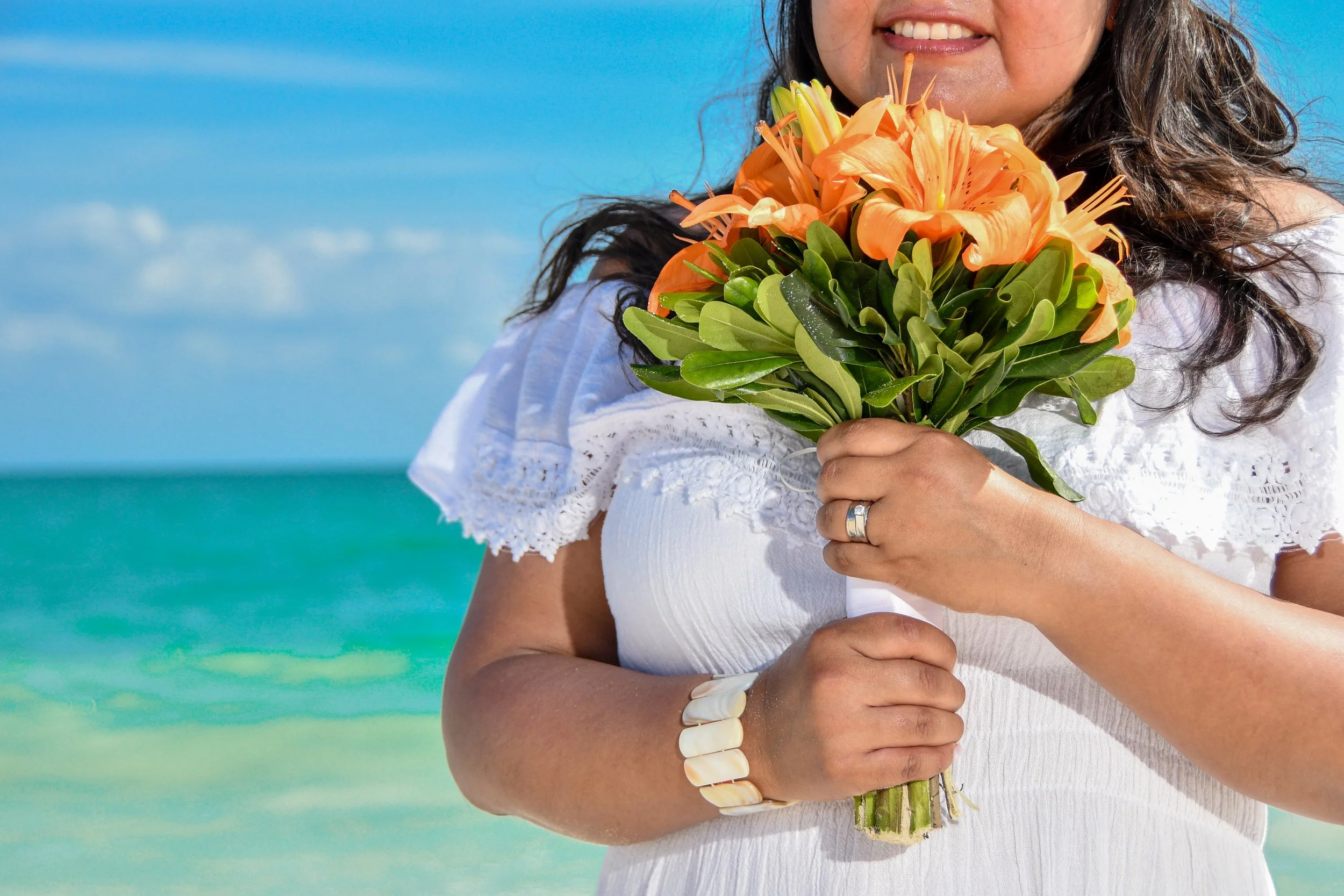 A woman holding a bouquet of orange flowers on a beach with clear blue sky and ocean in the background.