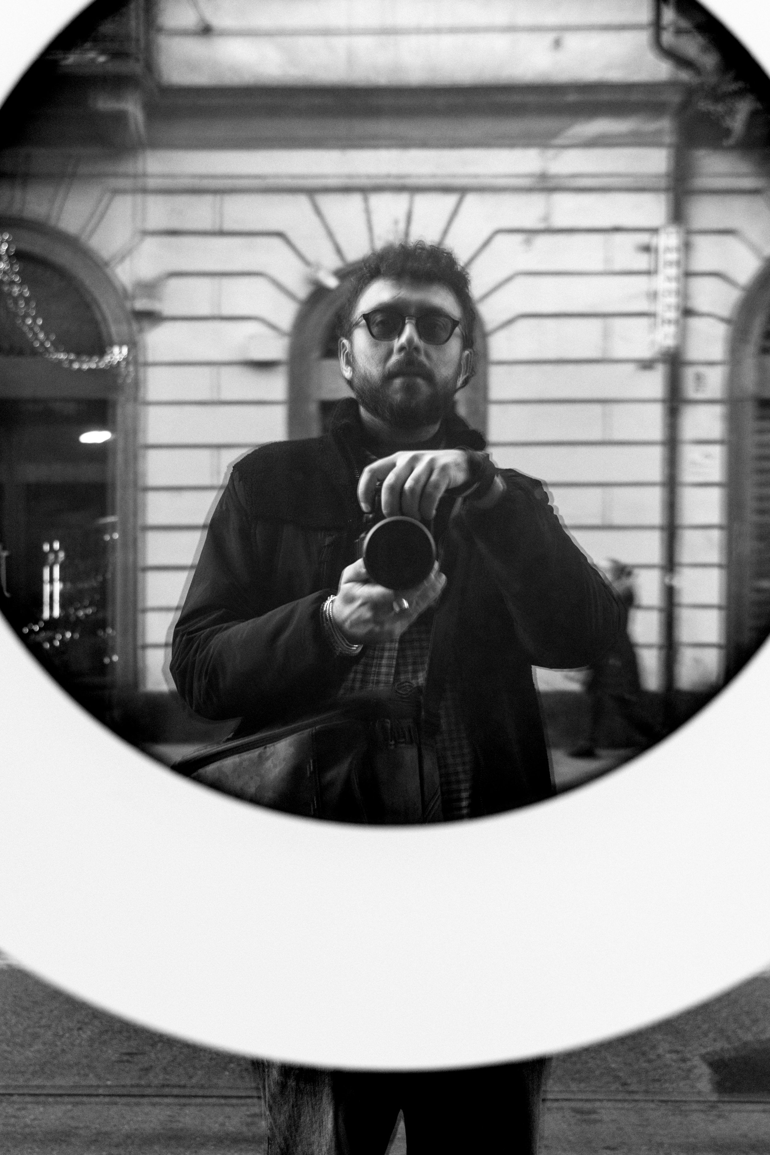 A man with glasses holding a camera taking a selfie is reflected in a mirror. Background includes a building with architectural details and a person walking by.