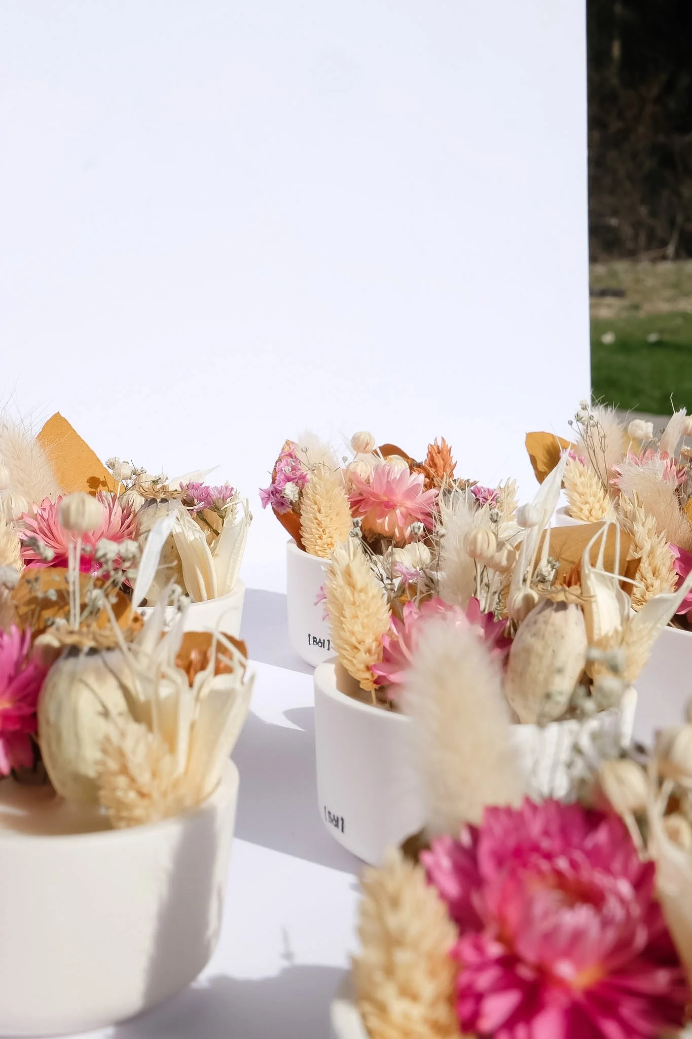 Petits arrangements floraux avec des fleurs séchées roses, blanches et beiges dans des contenants blancs, en plein air.