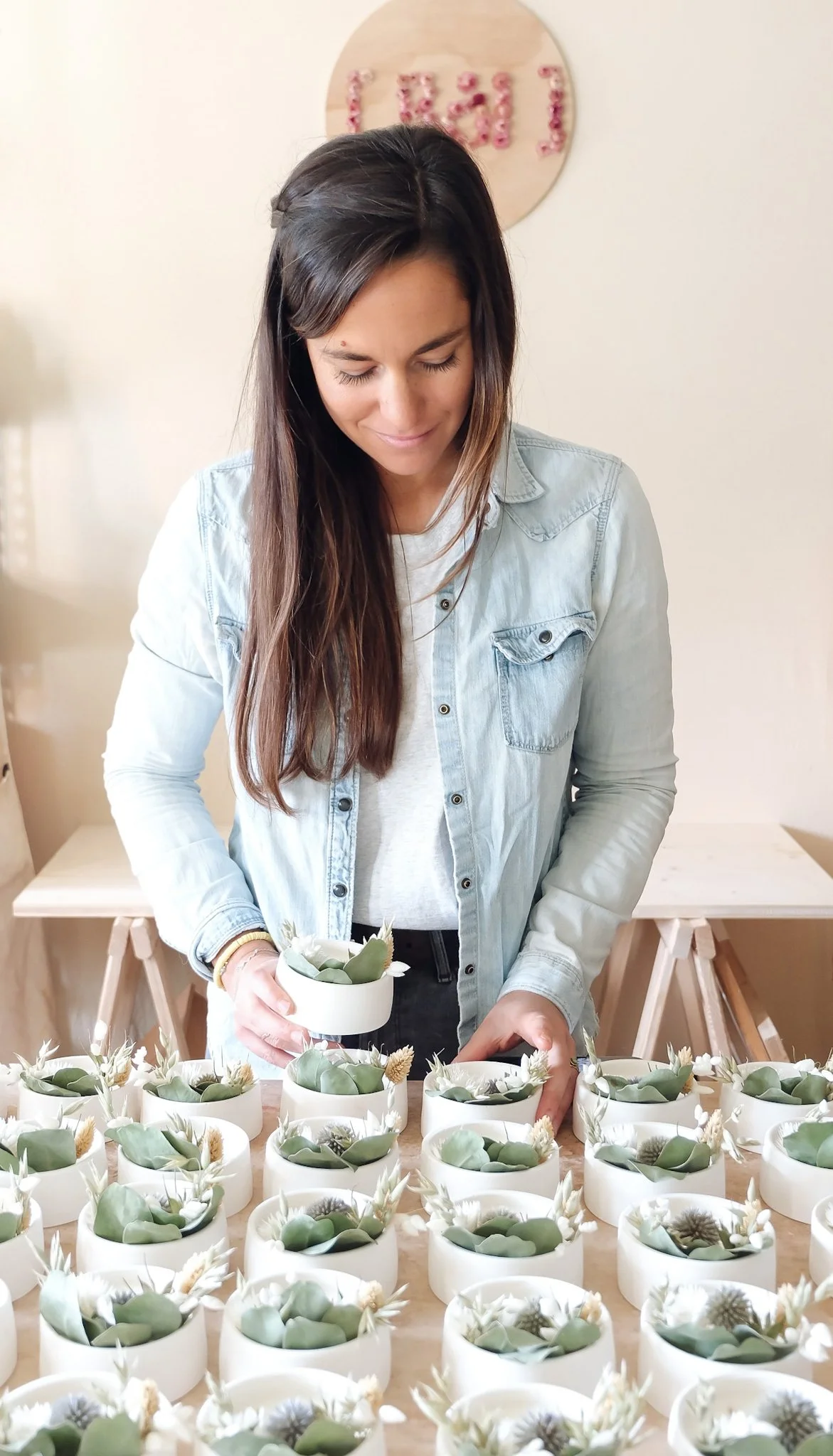 Une femme avec des cheveux longs et bruns, portant une veste en jean claire, arrange des plantes en pot blanches avec des fleurs blanches et de petites plantes, sur une table en bois, dans un intérieur lumineux.