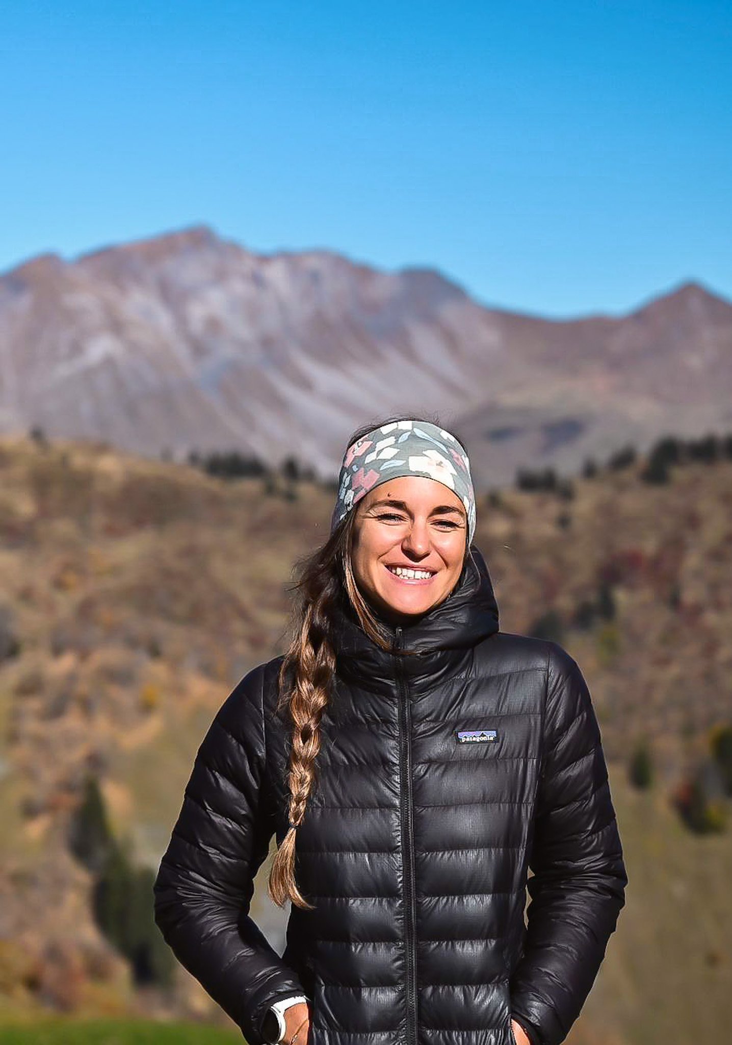 Femme souriante en veste noire, portant un bandeau à motifs, en plein air avec des montagnes en arrière-plan.