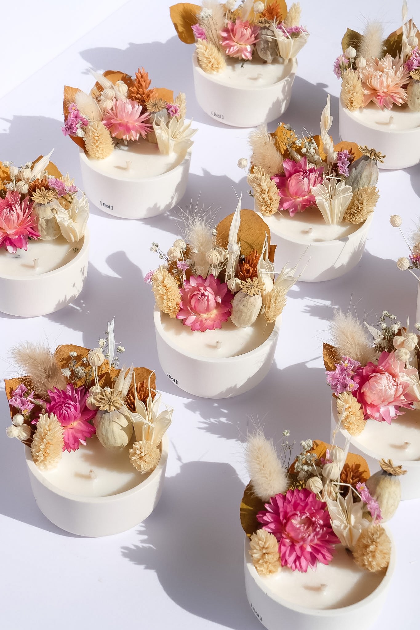 Bougies décoratives avec fleurs séchées roses, blanches et beiges, disposées dans des contenants ronds blancs, sur une surface blanche.