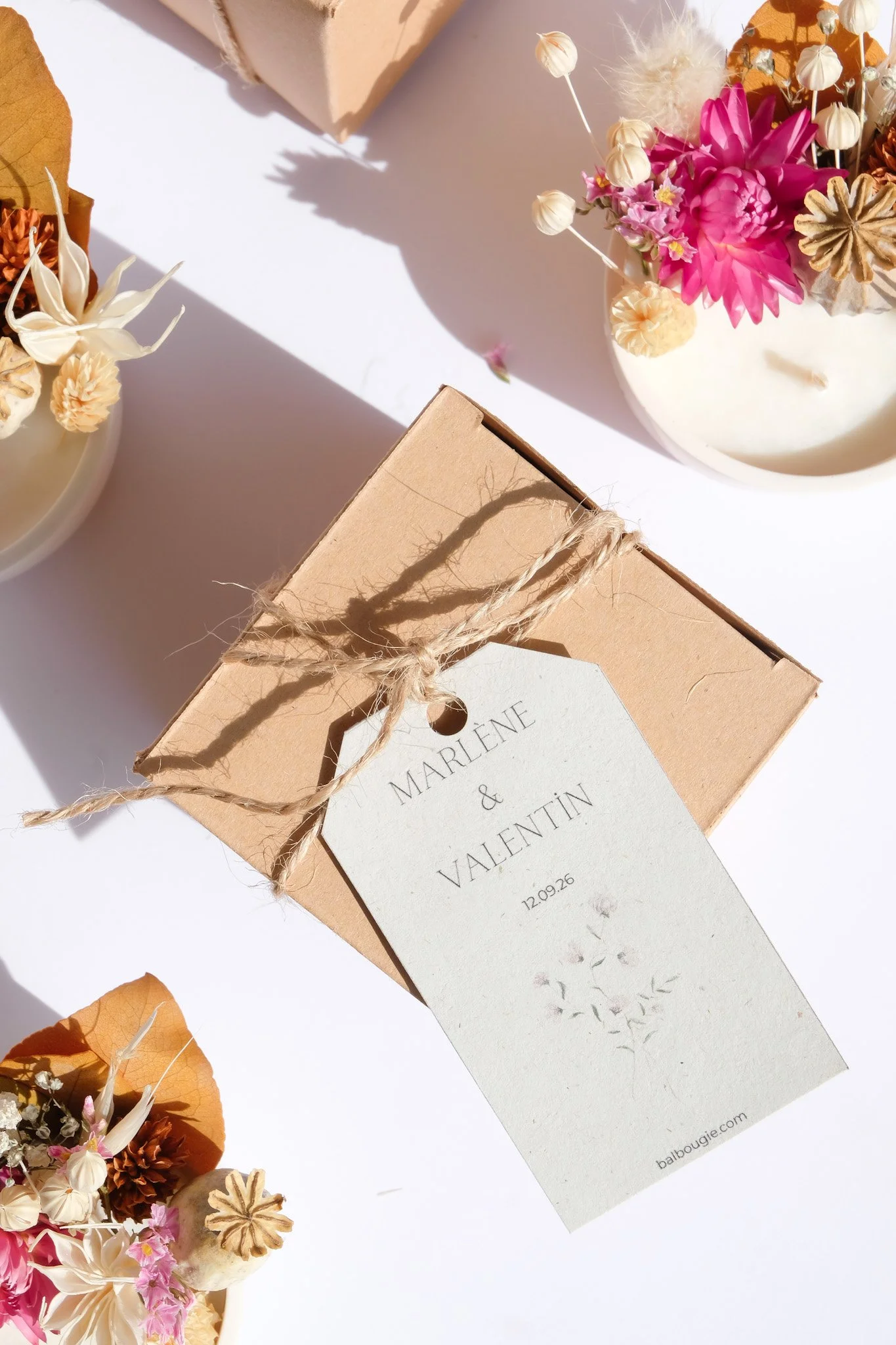 Une petite boîte en carton beige attachée avec une corde, avec une étiquette portant le nom Marlené & Valentin, et une date 12.09.26. La boîte est entourée de bouquets de fleurs séchées dans des vases blancs, placés sur une surface blanche, avec des ombres de lumière naturelle.