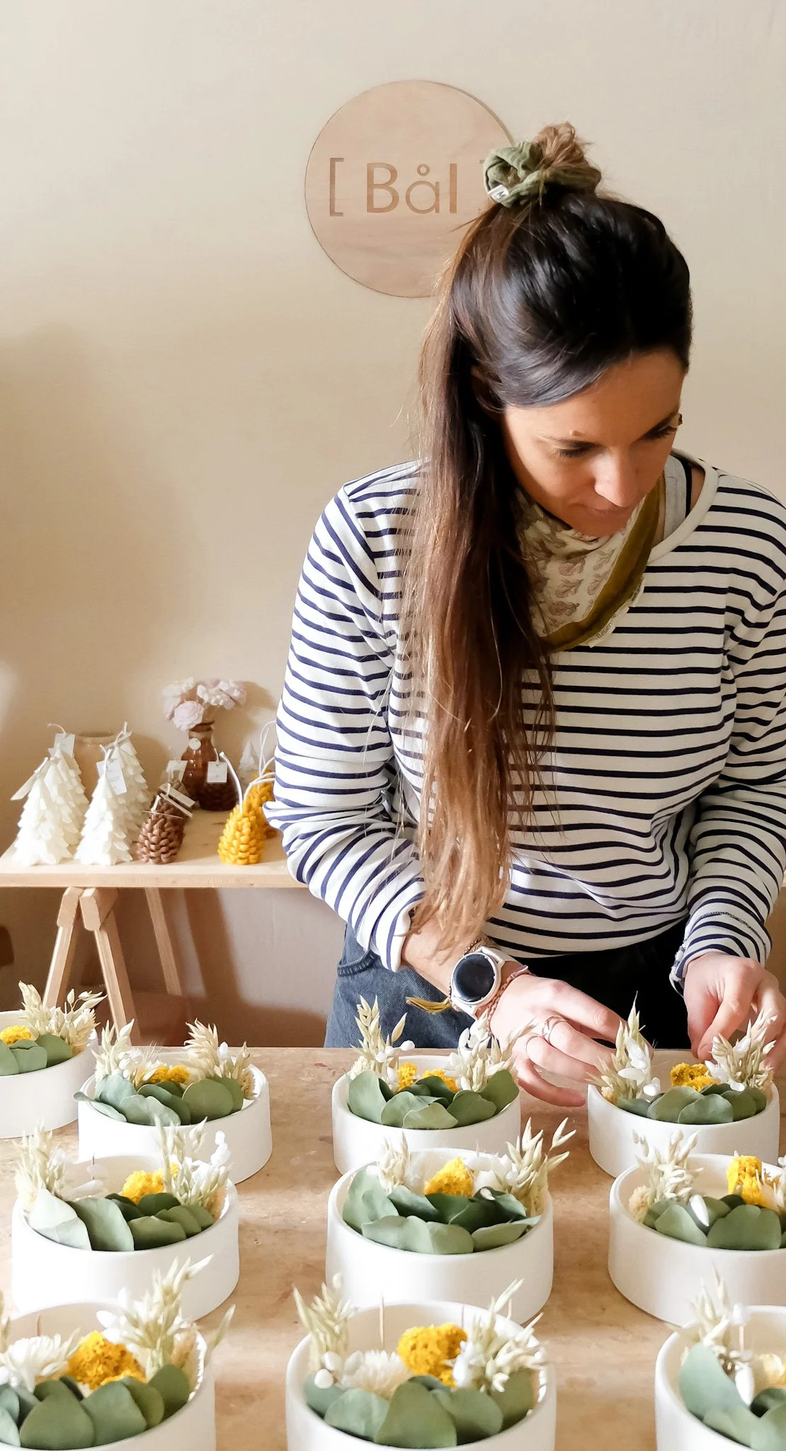 Une femme prépare des arrangements floraux avec des fleurs blanches et jaunes dans des boîtes blanches sur un comptoir en bois, dans un intérieur décoré avec des éléments de style naturel.