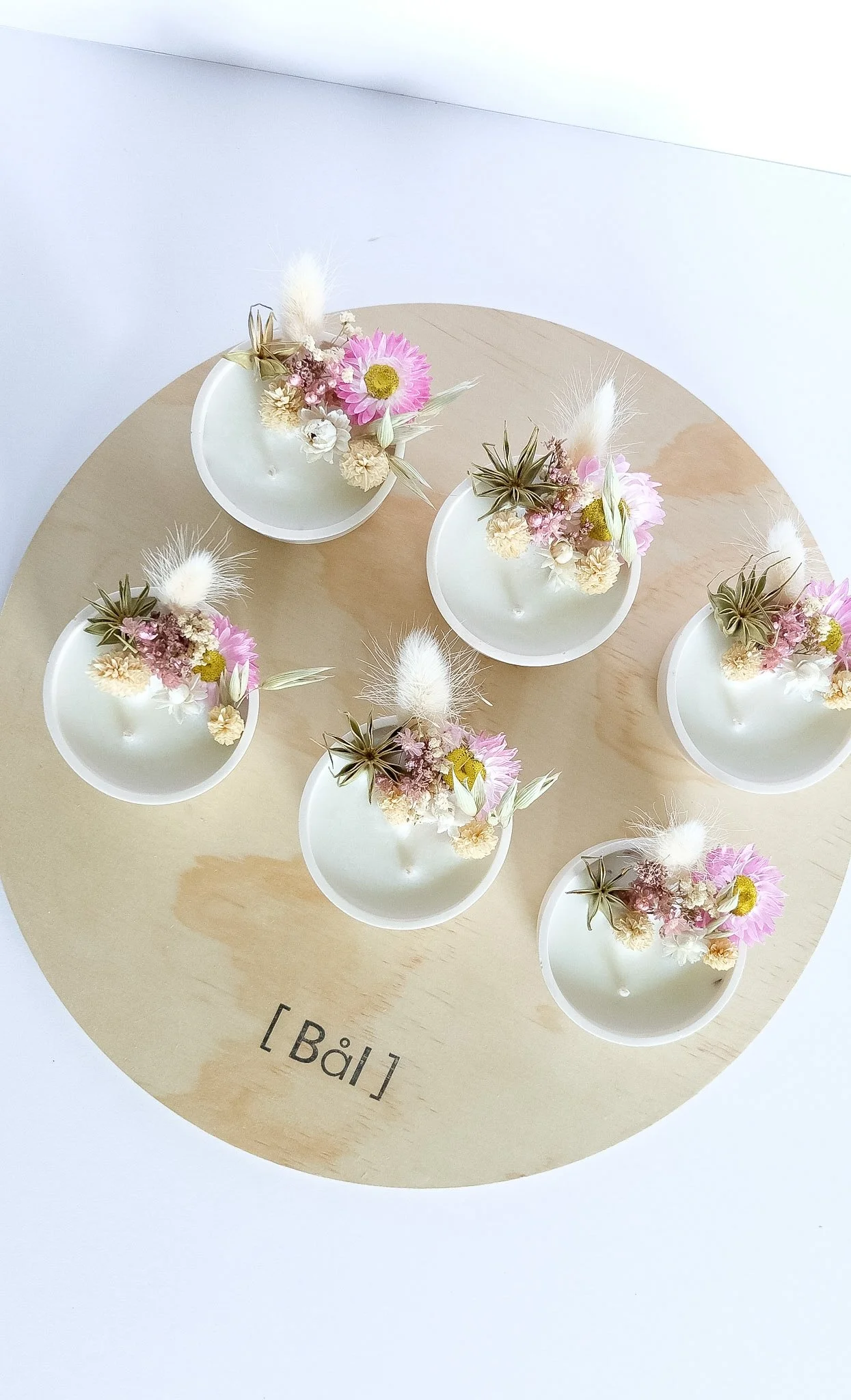 Six small white cups with floral arrangements, placed on a round wooden surface labeled '[Bâl]'. The arrangements include pink daisies, dried grasses, and other dried flowers.