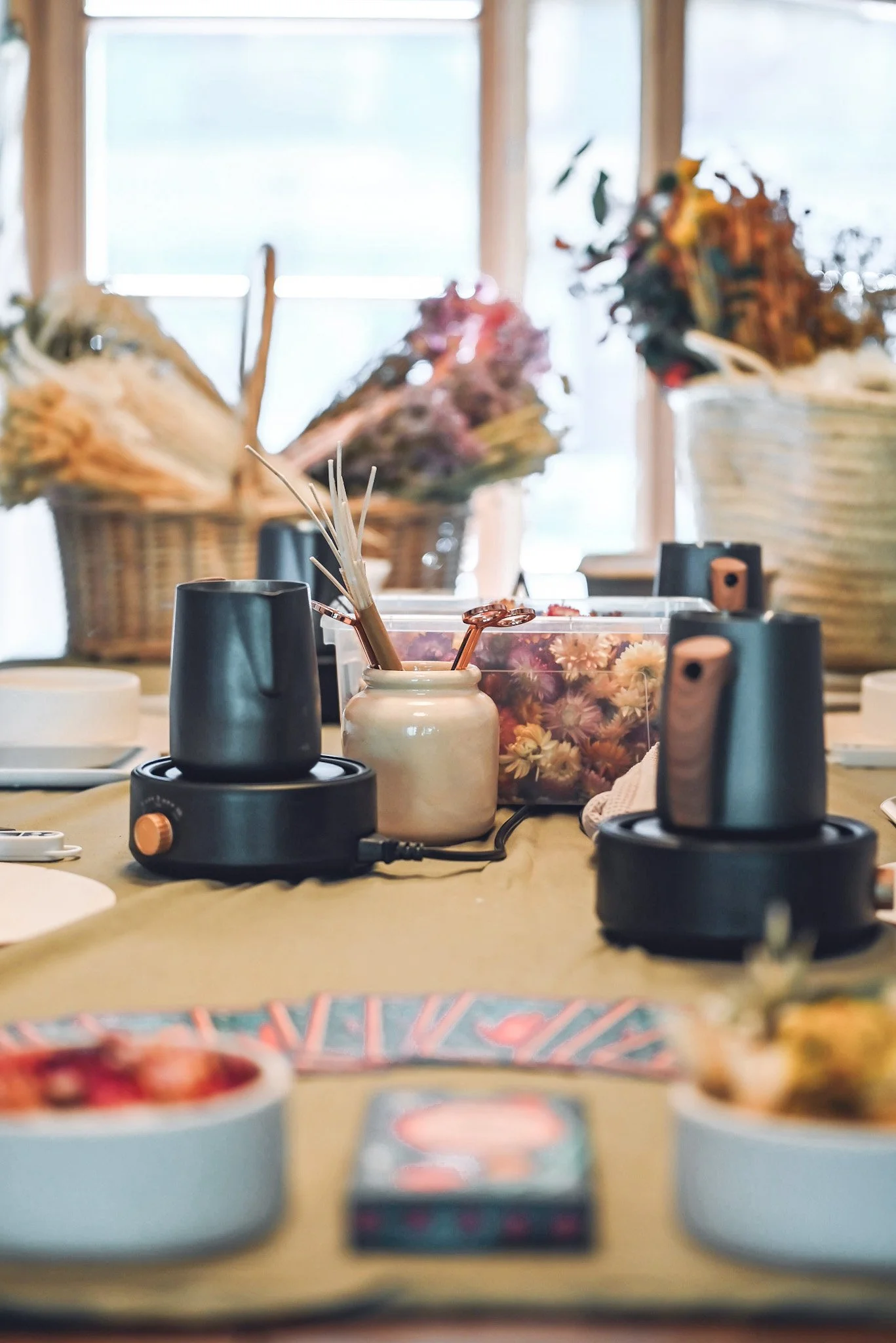 Table de dégustation avec deux cafetières électriques, un vase avec des brins de paille, et des bouquets de fleurs séchées en arrière-plan.