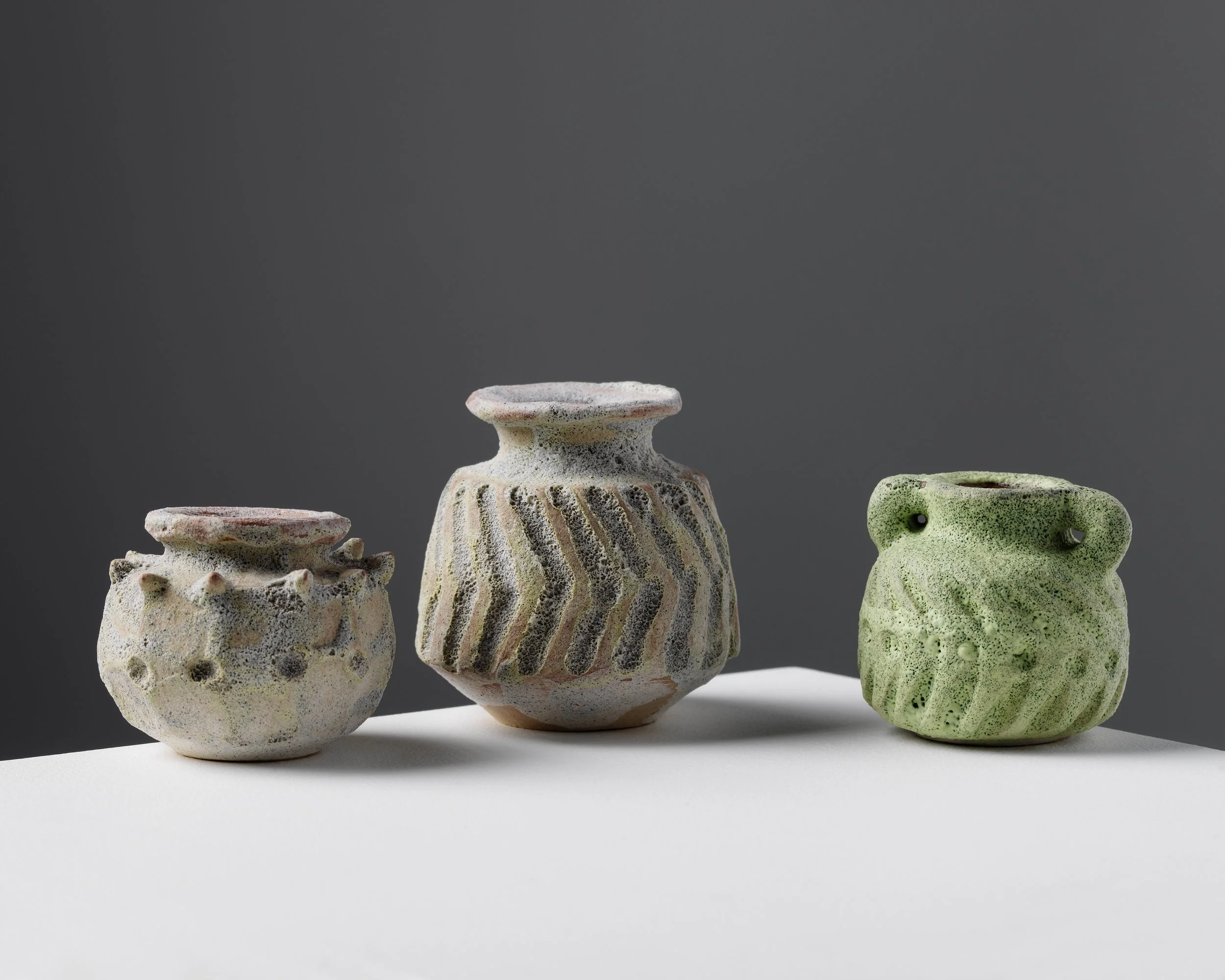 Three ceramic vases with textured and patterned surfaces, placed on a white surface against a dark gray background.