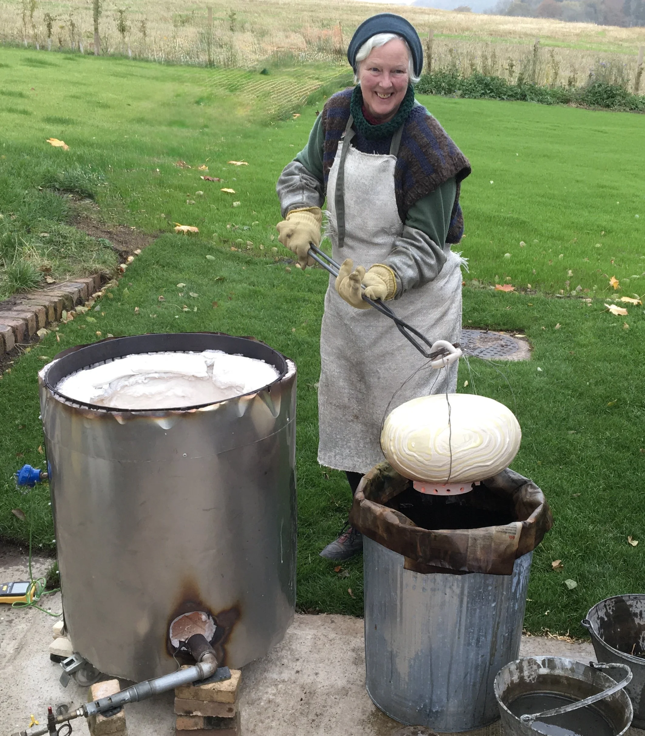 An elderly woman with white hair and a blue hat is outdoors, holding tongs and working with a hot kiln or furnace, shaping a large, light-colored glass or ceramic object. She is smiling and wearing a gray apron, gloves, and layered clothing. The background shows a grassy yard with fall leaves and a field.