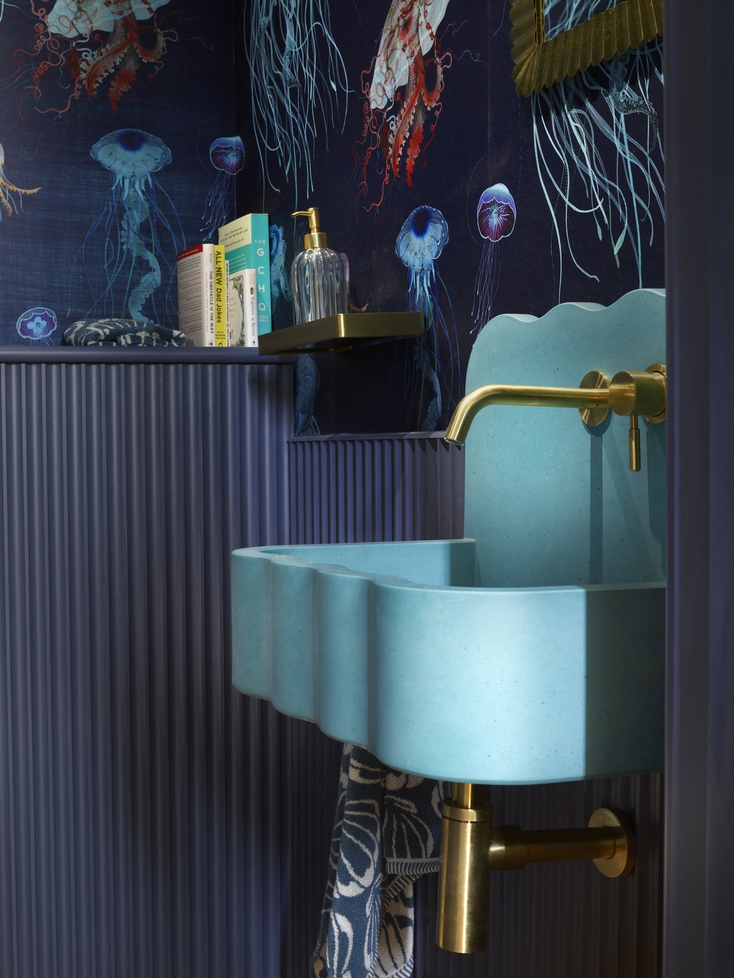 A small bathroom with a blue-green sink, gold fixtures, a dark blue fluted wall, and a dark blue wallpaper featuring jellyfish and ocean scenes. There are books and a soap dispenser on a small wall shelf above the sink.