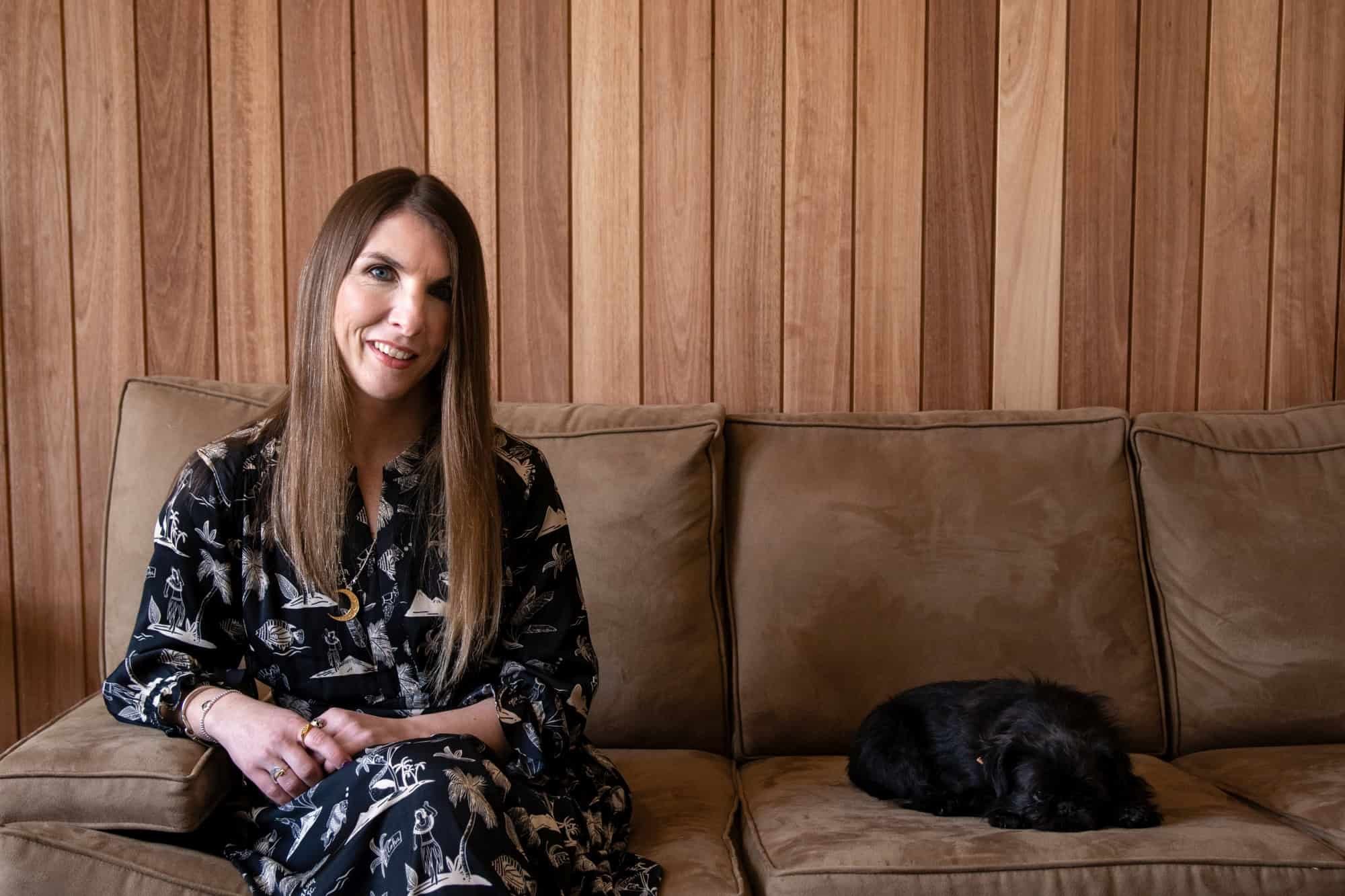 A woman, Katy Raywood, interior designer, with long straight brown hair and a black patterned dress sitting on a tan couch next to a sleeping black dog, with wooden paneling on the wall behind them.