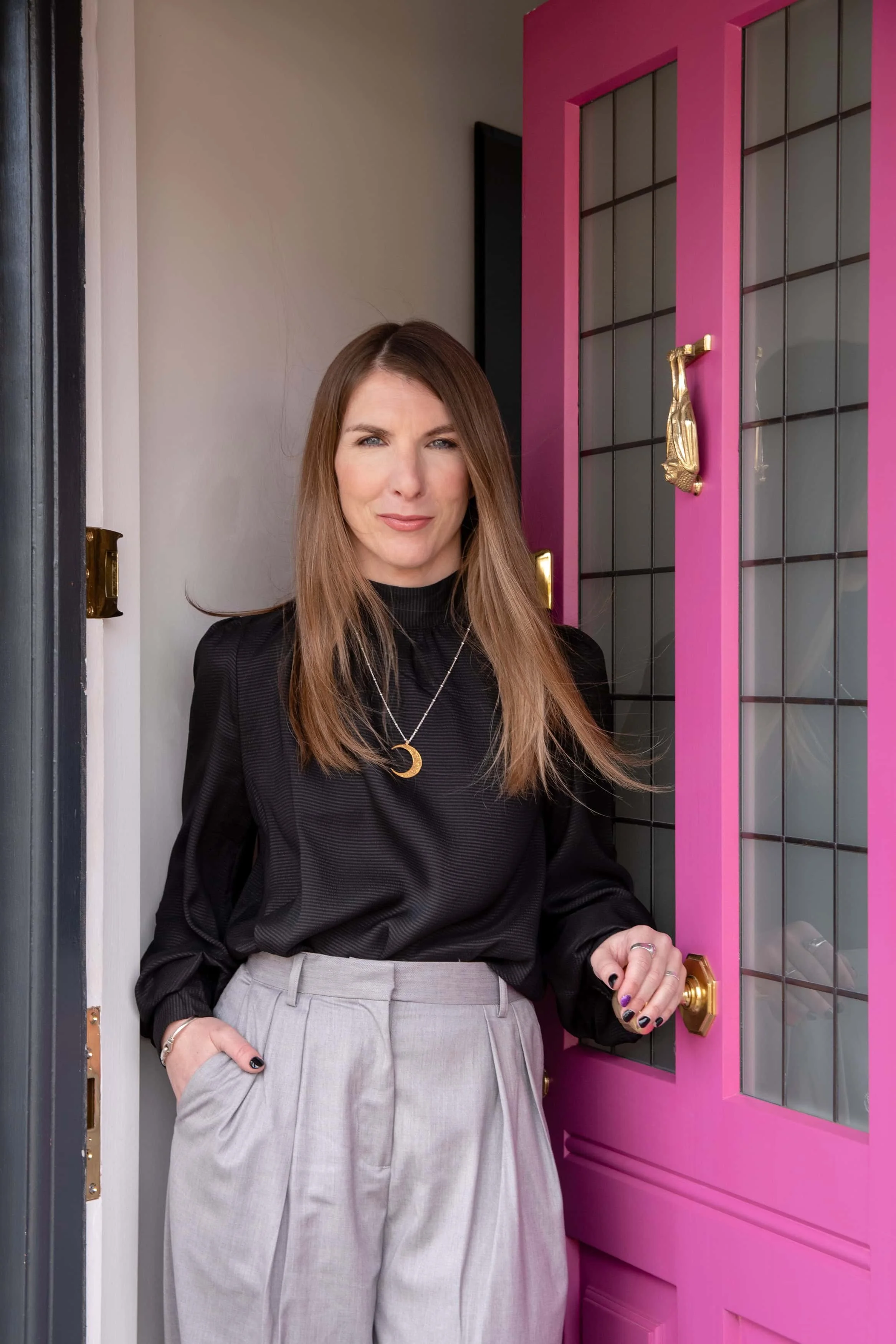 A woman with long, light brown hair, wearing a black top, gray high-waisted trousers, and jewelry, standing in a doorway with a black frame and a pink door with glass panes and gold hardware, smiling slightly.