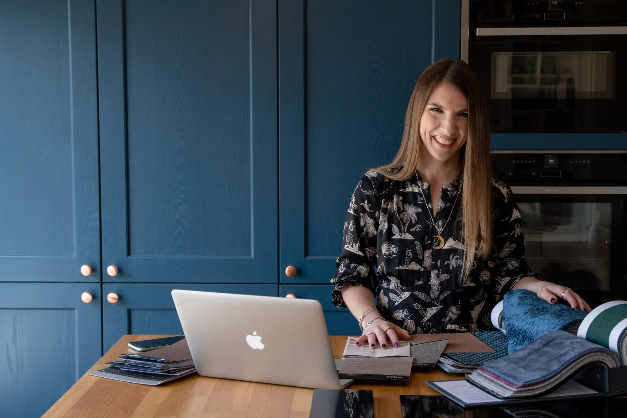 A woman with long brown hair smiling in a modern kitchen, standing behind a wooden table with fabric samples, a silver MacBook, and a smartphone.