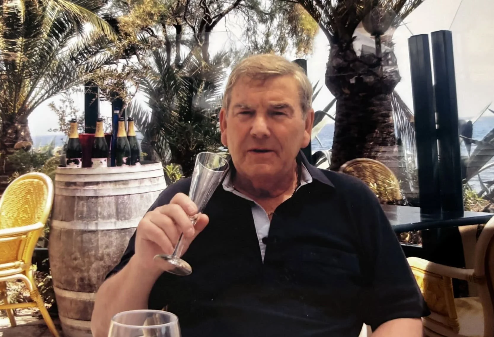 A man with gray hair wearing a black shirt, sitting outdoors, holding a champagne flute, with tropical trees and a barrel with bottles in the background.