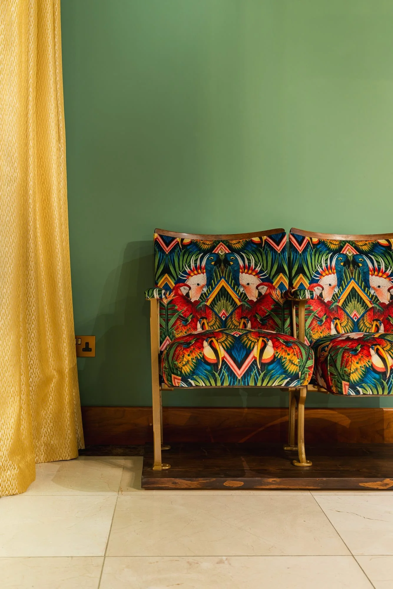 Colorful vintage cinema seats with a vibrant bird and foliage pattern, placed against a green wall with a yellow curtain to the left.