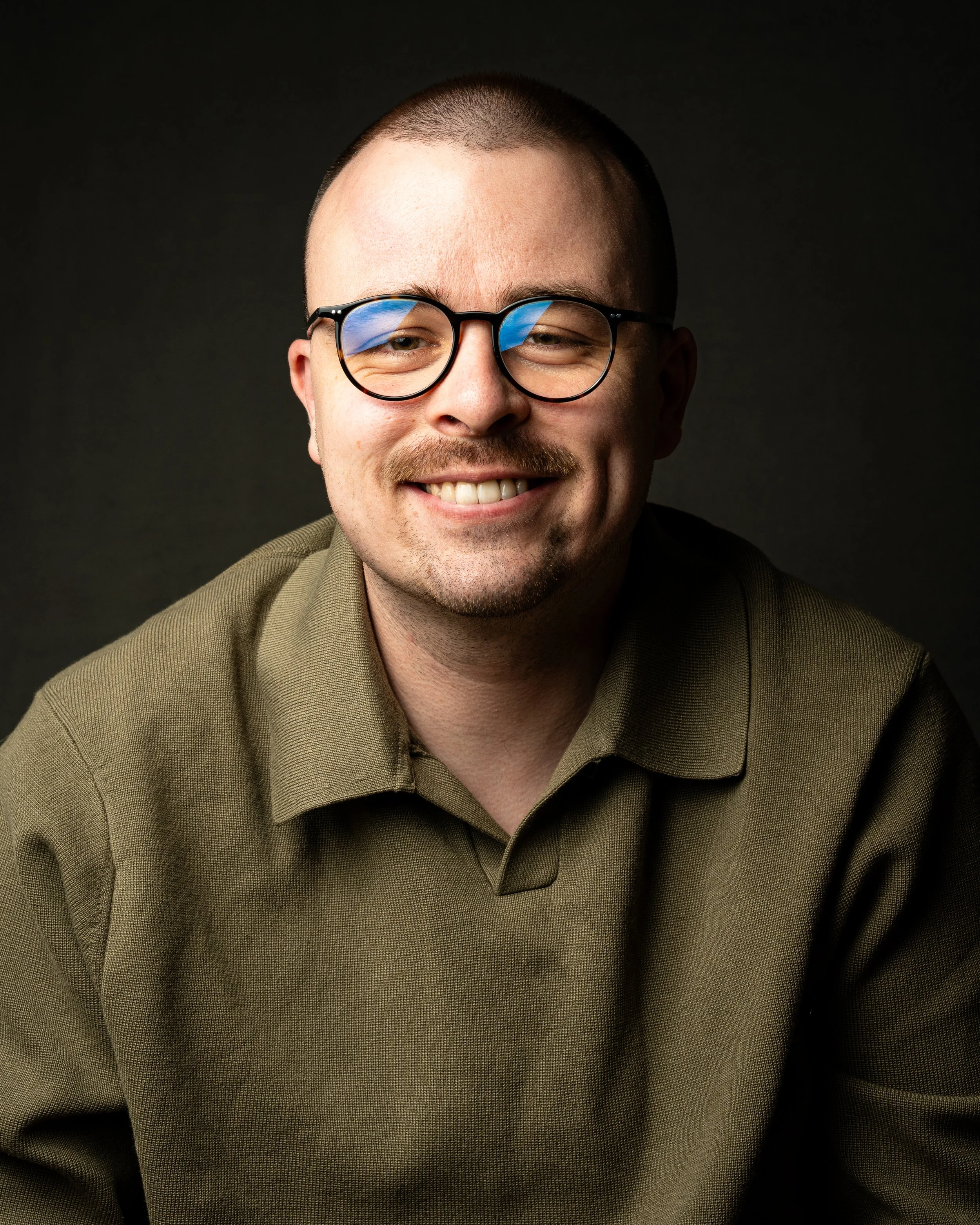 Porträt eines Mannes mit Brille, kurzem Haar, Bart und einem freundlichen Lächeln, trägt ein olivgrünes Hemd, vor dunklem Hintergrund.