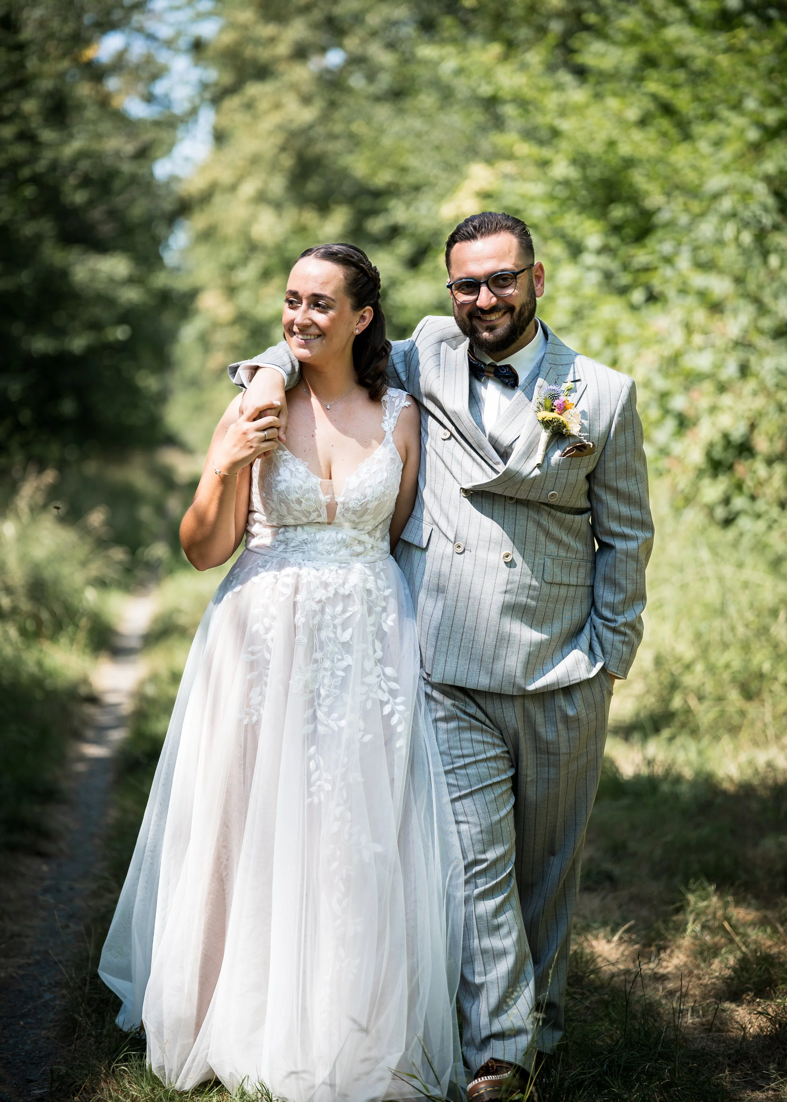Ein glückliches Brautpaar läuft durch einen grünen Park, die Braut trägt ein weißes, spitz zulaufendes Brautkleid mit Blumenapplikationen, der Bräutigam trägt einen grauen, gestreiften Anzug mit Fliege und Brille. Das Paar lächelt fröhlich, umgeben v