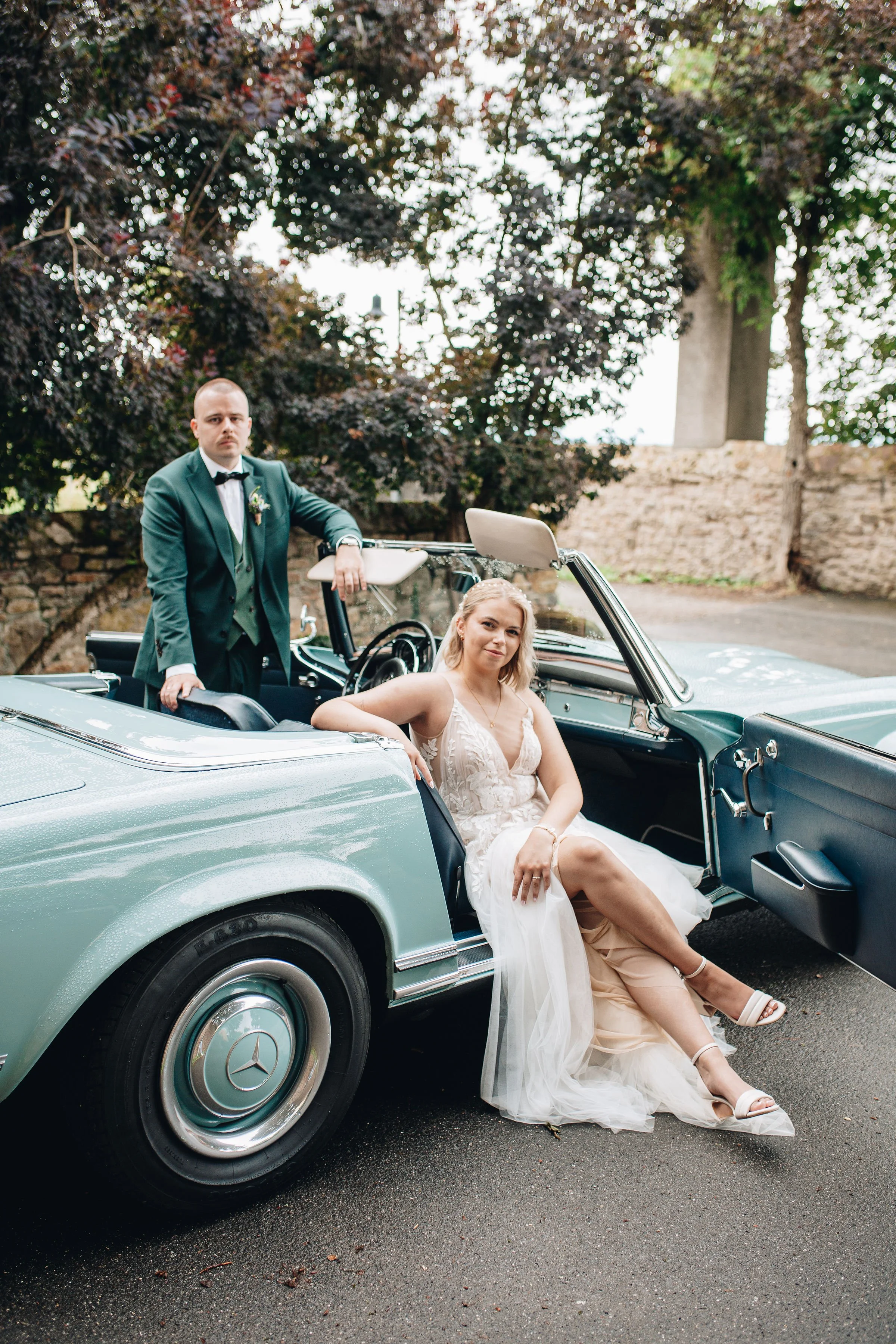 Ein Brautpaar posiert in einem Oldtimer-Cabrio auf einem Straßenabschnitt mit Bäumen im Hintergrund.