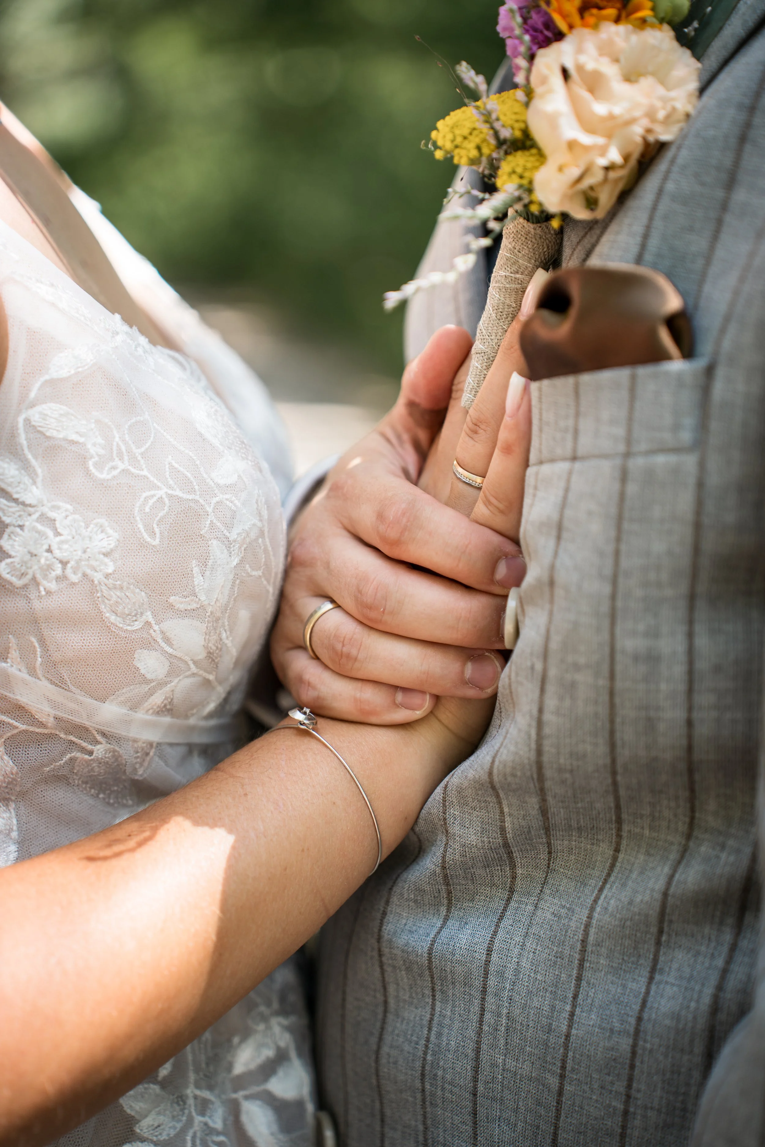 Nahaufnahme eines Paares an ihrer Hochzeit, nur die Hände und ein Teil des Brautkleids sowie des Anzugs sind sichtbar. Die Braut trägt ein Ring und ein Armband, der Bräutigam trägt einen Ring. Die Braut hält einen kleinen Blumenstrauß. Die Szene ist 
