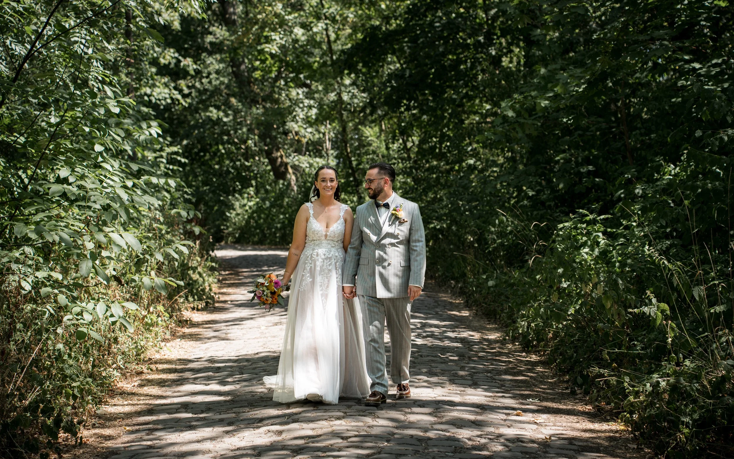 Ein Brautpaar, eine Frau in einem weißen Hochzeitskleid und ein Mann in einem grauen Anzug, spazieren Hand in Hand durch einen von Bäumen umgebenen Waldweg. Die Frau hält einen bunten Blumenstrauß in der Hand und beide lächeln freundlich.
