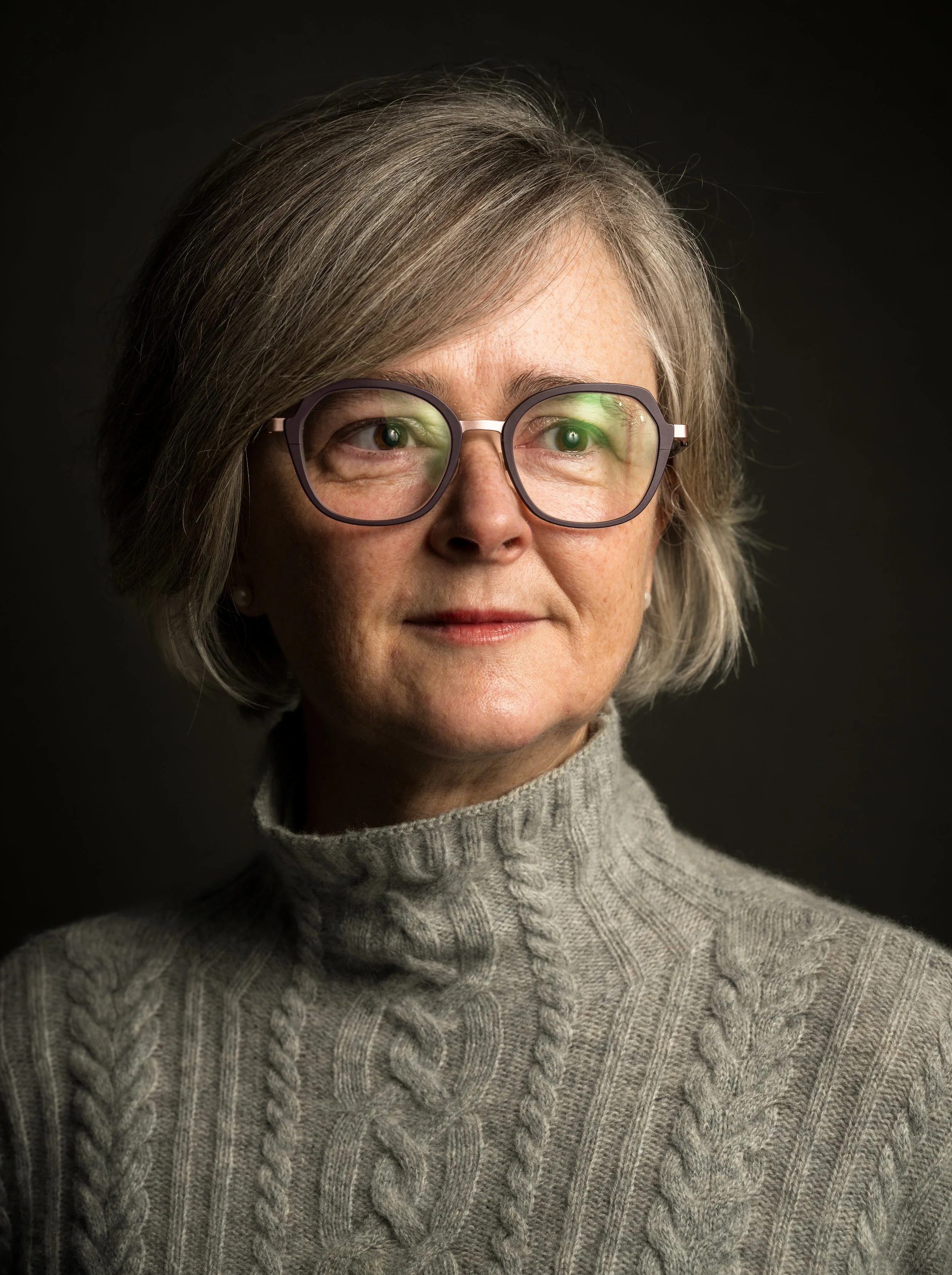 Nahaufnahme einer älteren Frau mit grauen Haaren, Brille und grauem Pullover vor schwarzem Hintergrund.