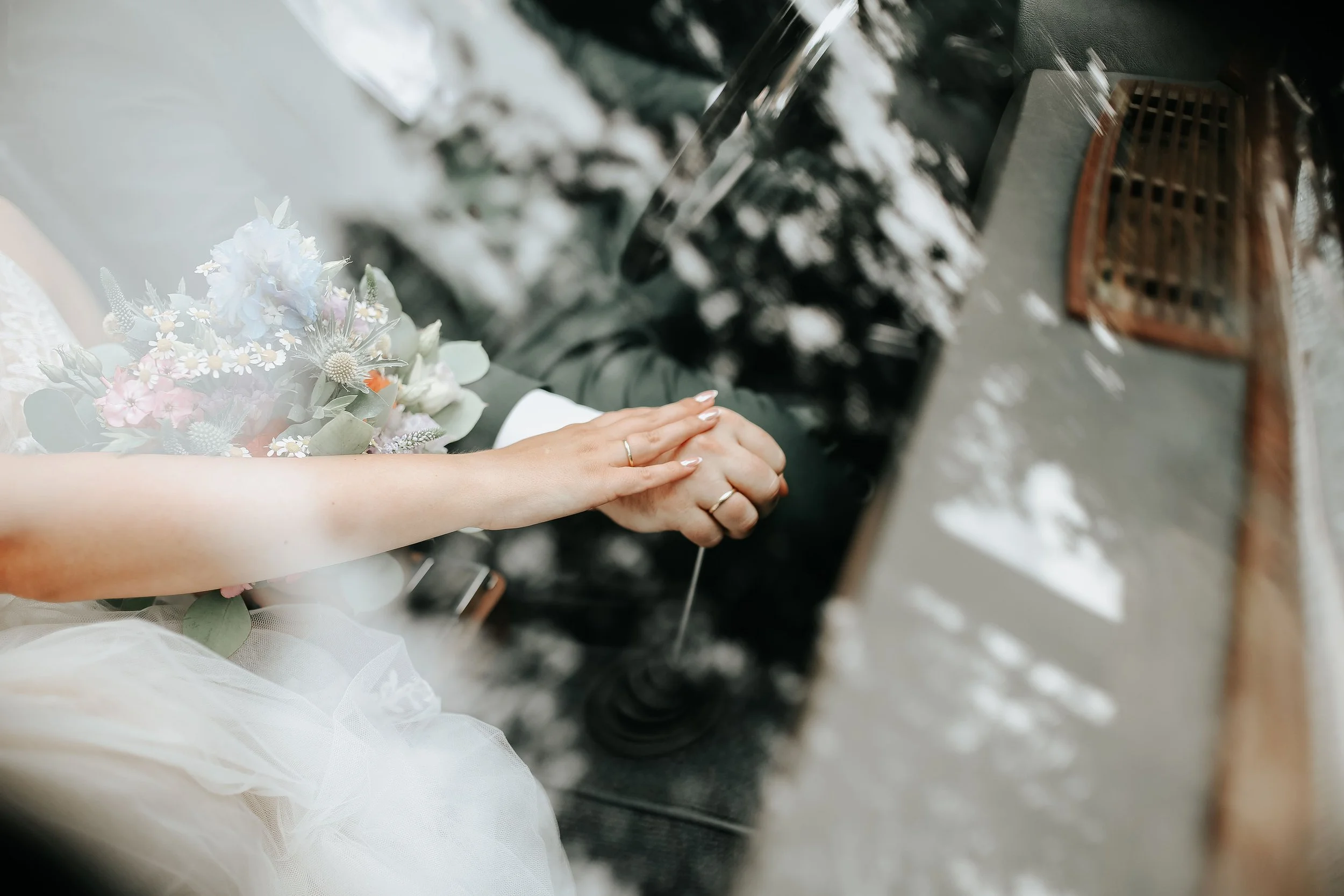 Nahaufnahme von den Händen eines Paares, die bei einer Hochzeit Händchen halten, mit einem Blumenstrauß und einer Kirchenbank im Hintergrund.