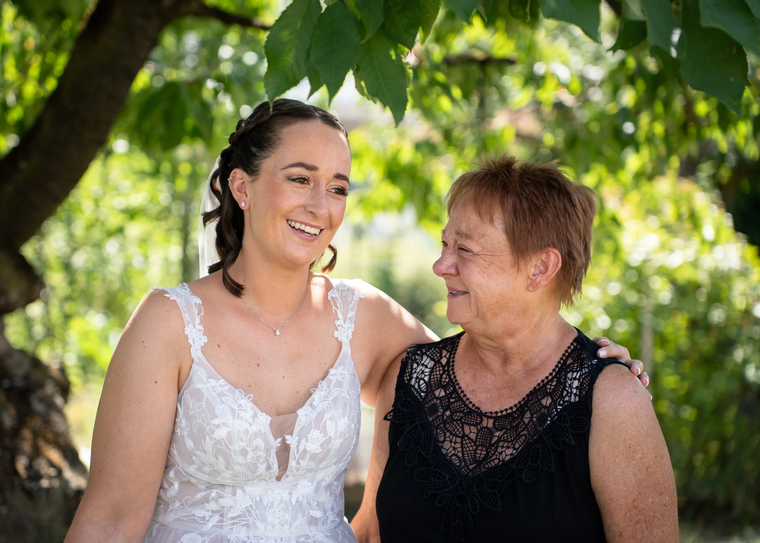 Zwei Frauen, eine junge Braut und eine ältere Frau, lächeln sich im Schatten eines Baumes an, umgeben von grünen Blättern, bei schönem Wetter im Freien.