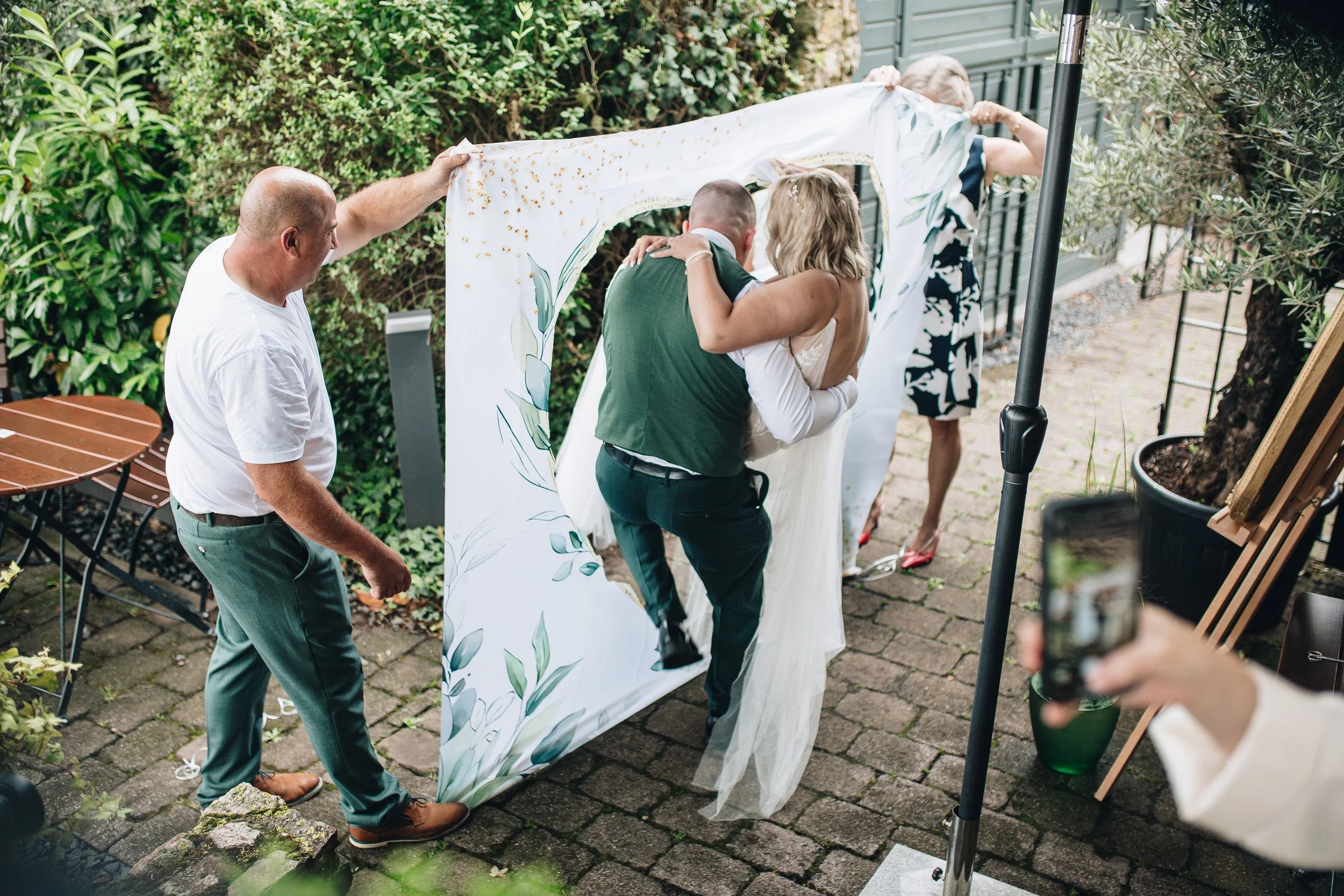 Ein Brautpaar wird beim Fotoshooting von vier Helfern mit einem weißen Stoffhintergrund umarmt, während sie sich umarmt.