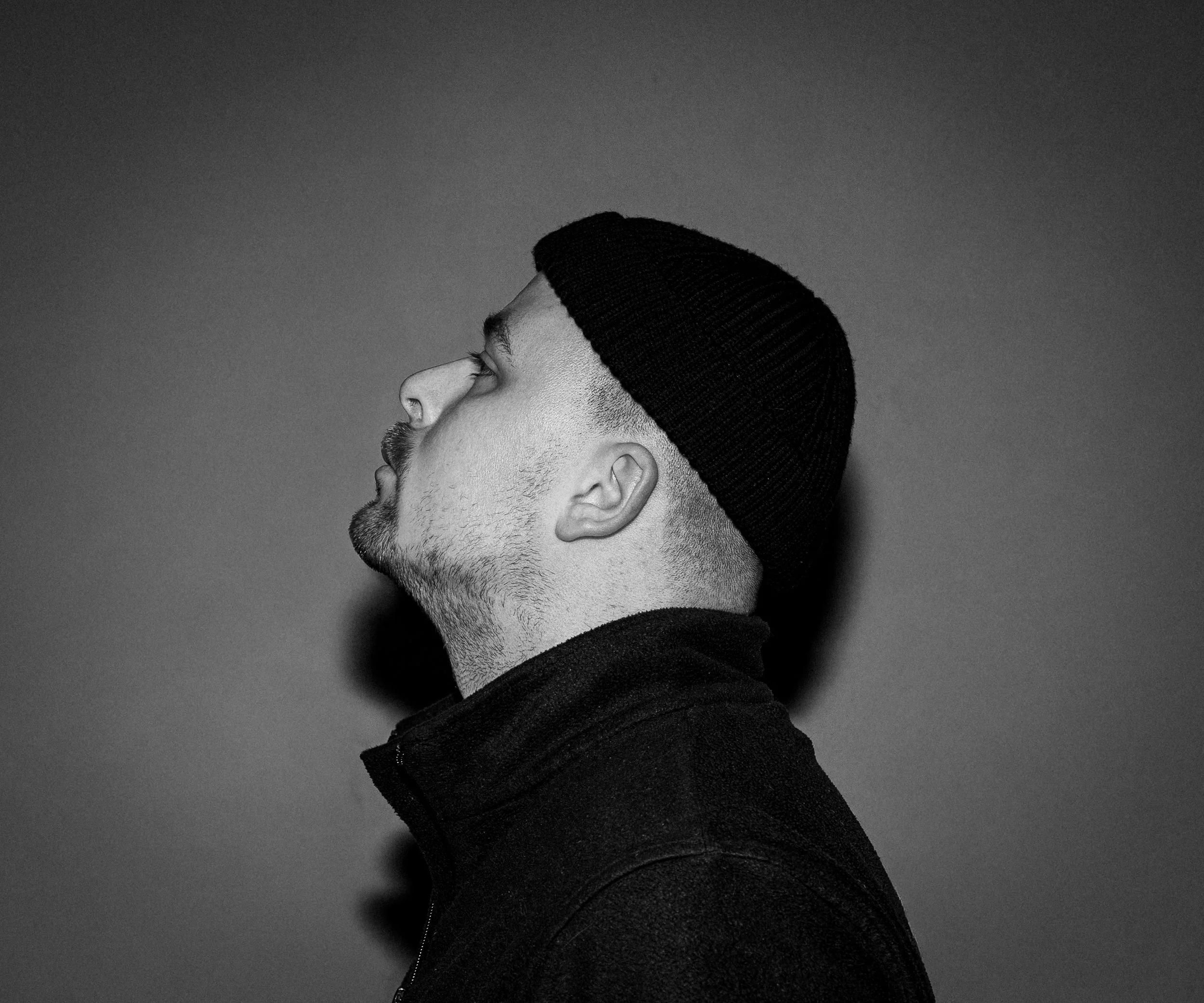 Porträt eines Mannes in Schwarz-Weiß, seitlich aufgenommen, trägt eine schwarze Mütze und einen schwarzen Hoodie.