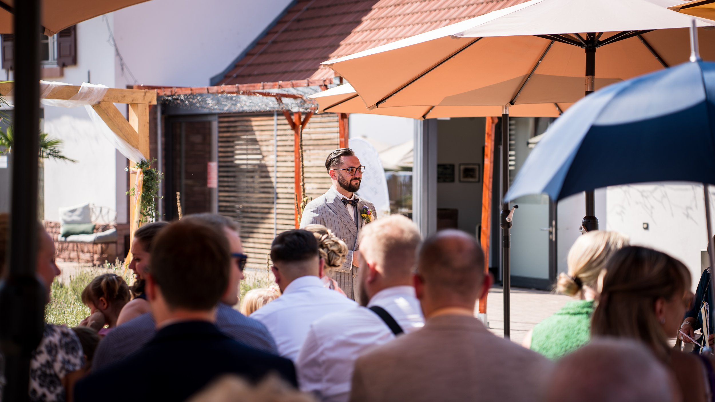 Mann trägt Anzug mit Krawatte und Brille, steht vor Publikum bei einer Veranstaltung im Freien unter Sonnenschirmen.
