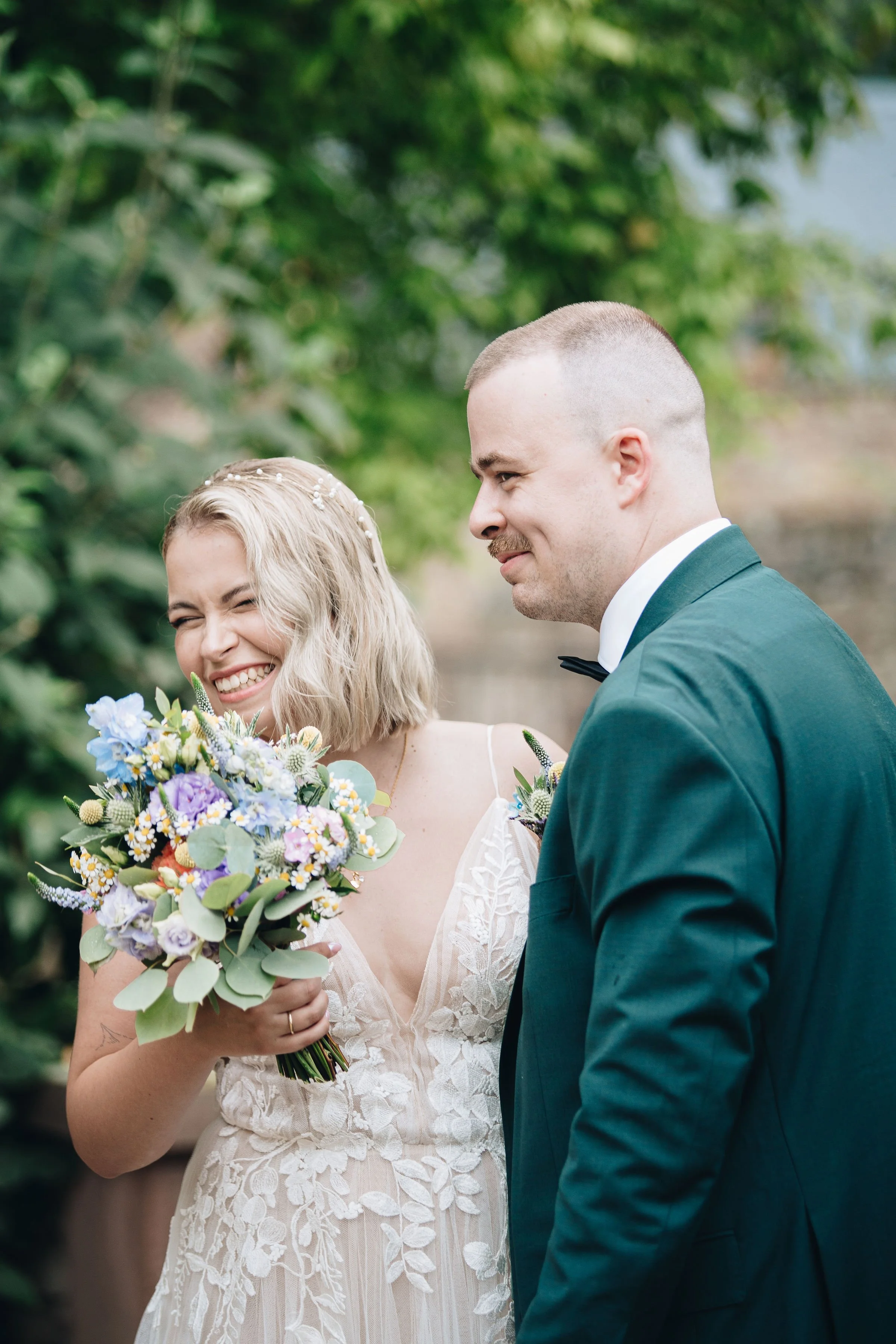 Ein lachendes Brautpaar bei ihrer Hochzeit im Freien, die Braut hält einen farbenfrohen Blumenstrauß, der Bräutigam trägt einen dunklen Anzug mit Fliege.