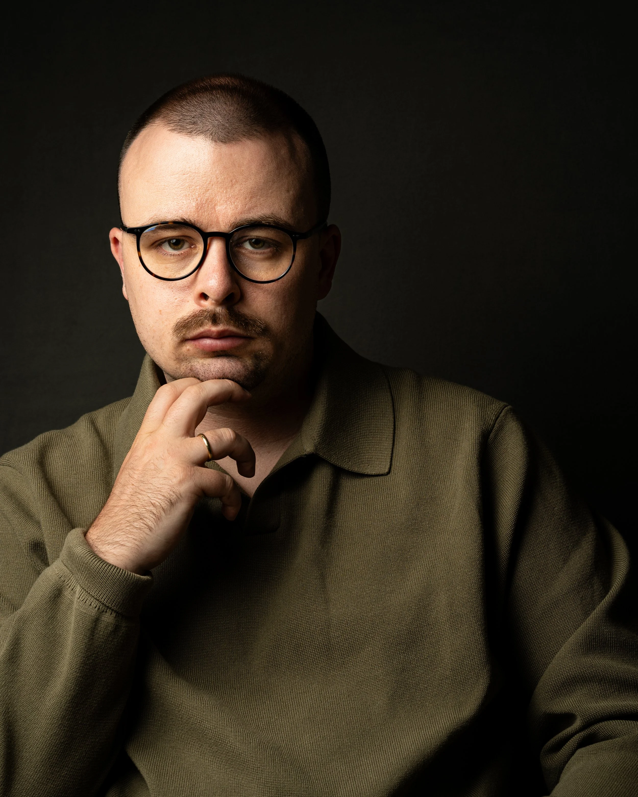 Nahaufnahme eines Mannes mit Brille, Bart und Kurzhaarschnitt, der nachdenklich die Hand an das Kinn legt, vor dunklem Hintergrund.