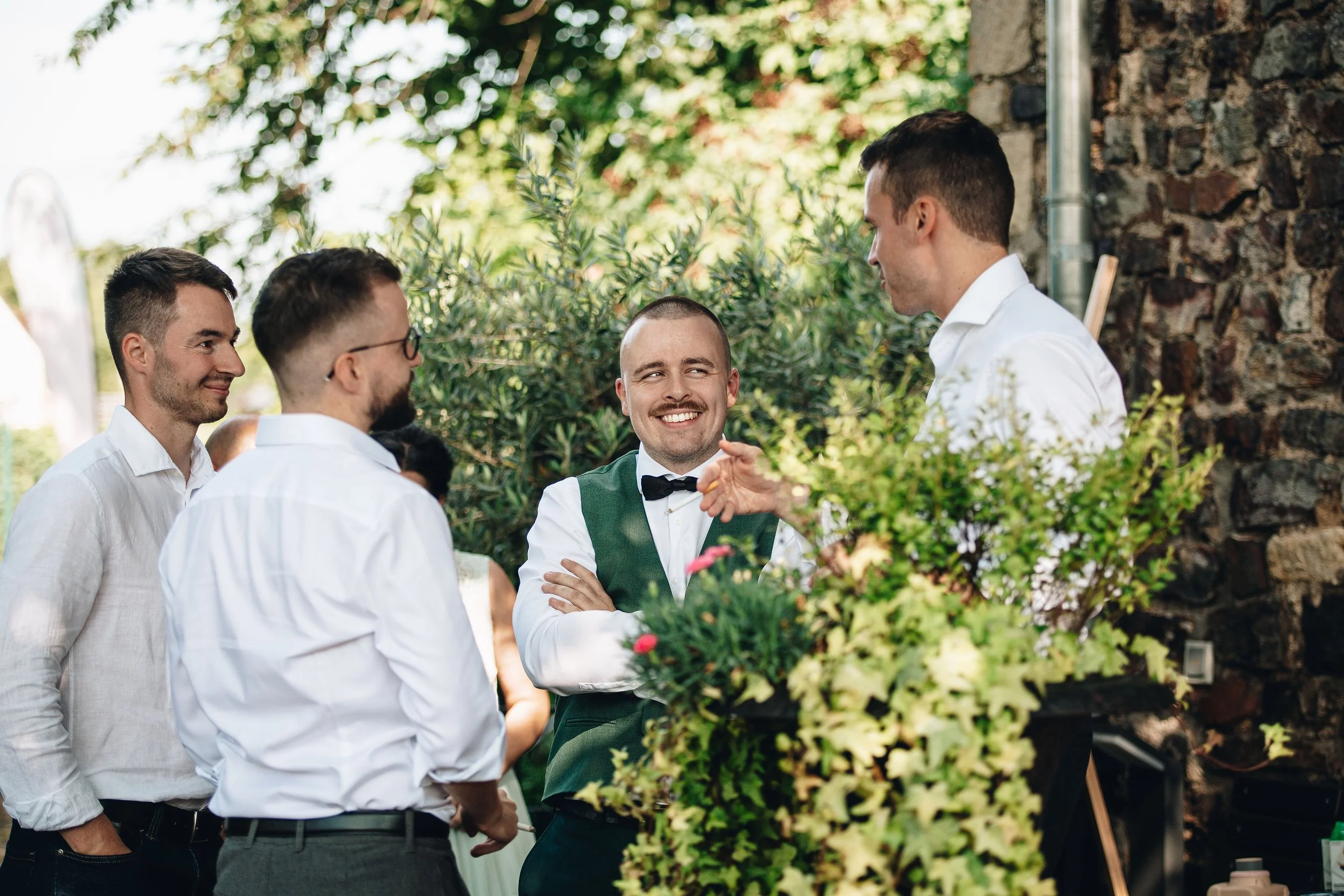Eine Gruppe von fünf jungen Männern, alle in formeller Kleidung, spricht und lacht im Freien vor einer Steinmauer. Es herrscht eine lockere und fröhliche Atmosphäre.