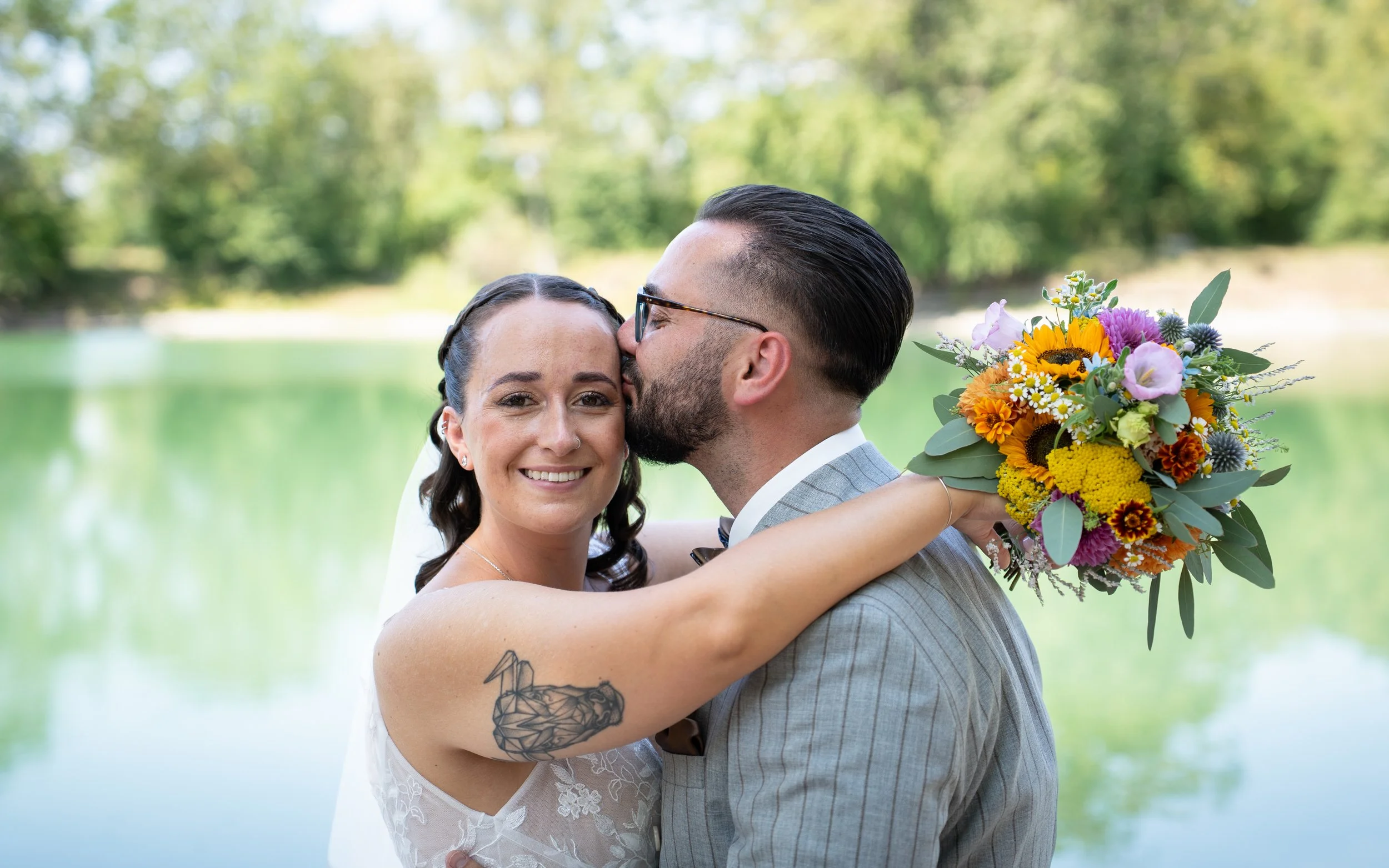 Ein glückliches Hochzeitspaar umarmt sich am Ufer eines Sees im Grünen, der Bräutigam küsst die Braut auf die Stirn, während sie in einem hellen Brautkleid und einem Anzug gekleidet sind, mit einem großen Blumenstrauß.