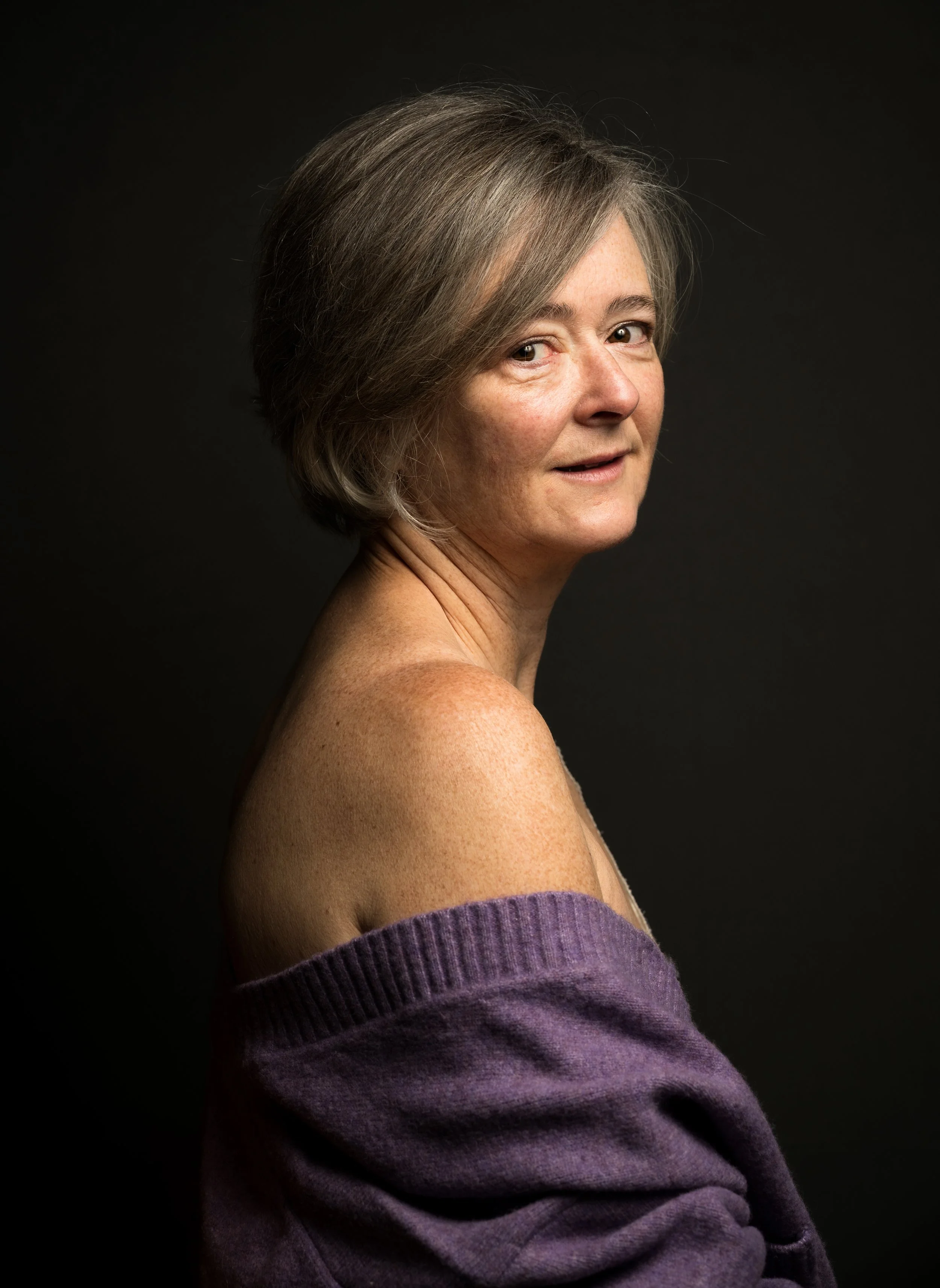 Porträt einer älteren Frau mit kurzen grauen Haaren, die eine lila Pullover trägt, vor schwarzem Hintergrund.