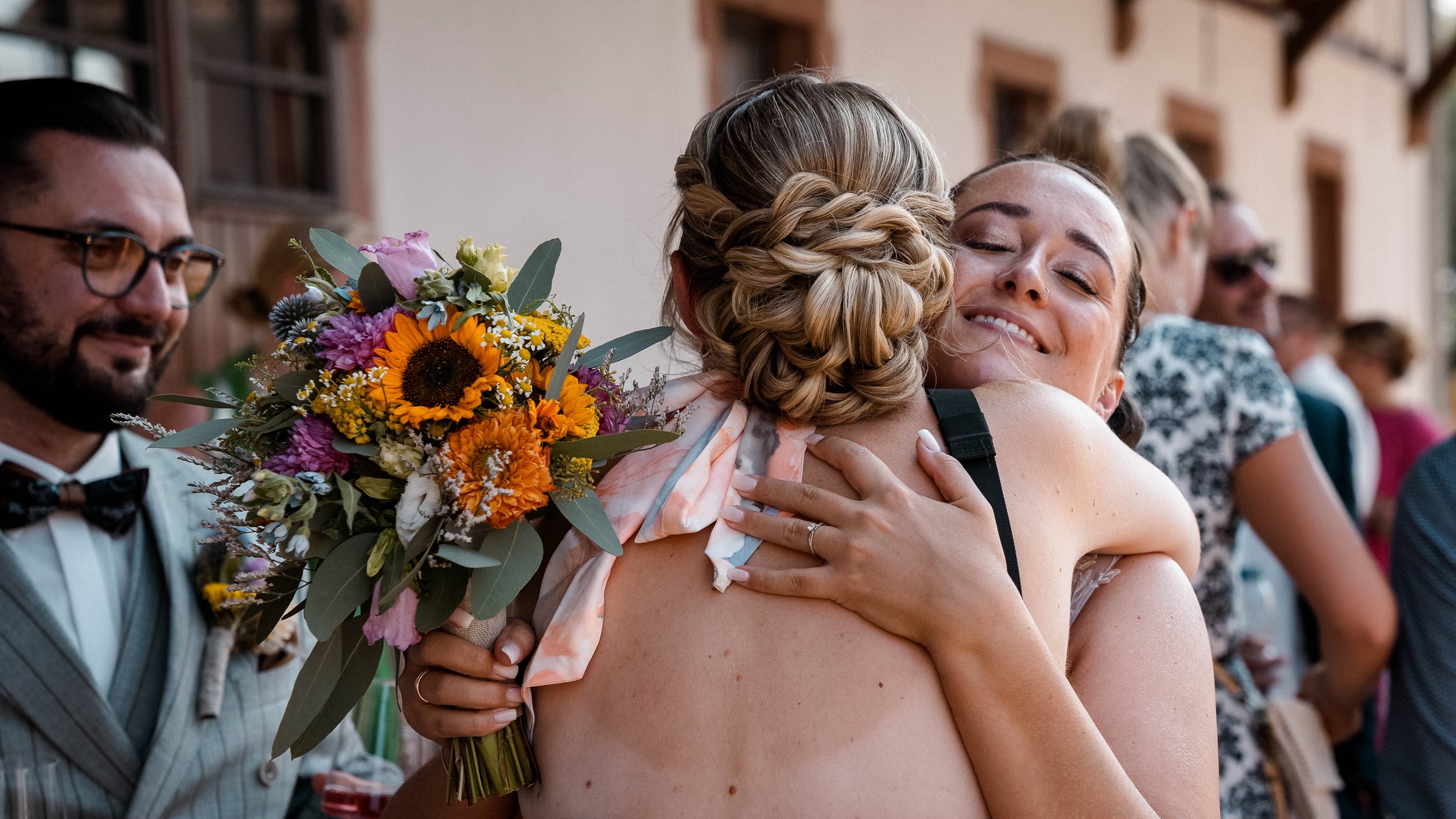 Zwei Frauen umarmen sich glücklich bei einer Hochzeit, eine hält einen bunten Blumenstrauß
