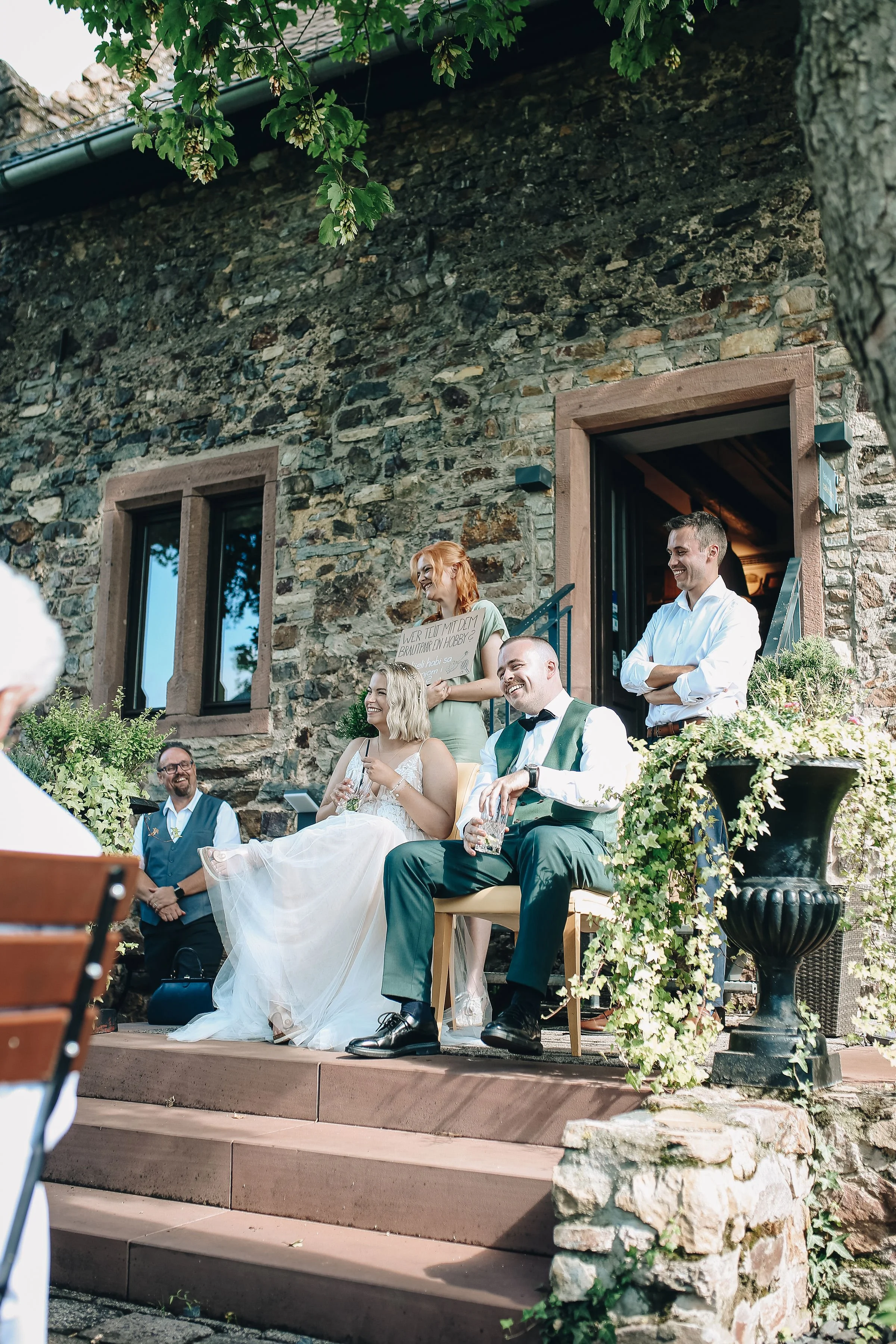 Eine Gruppe von Menschen bei einer Hochzeit im Freien, sitzend auf einer kleinen Treppe vor einer alten Steinmauer, darunter eine Braut in weißem Kleid, ein Bräutigam in Anzug mit Fliege, und andere Gäste, die lachen und sich unterhalten.