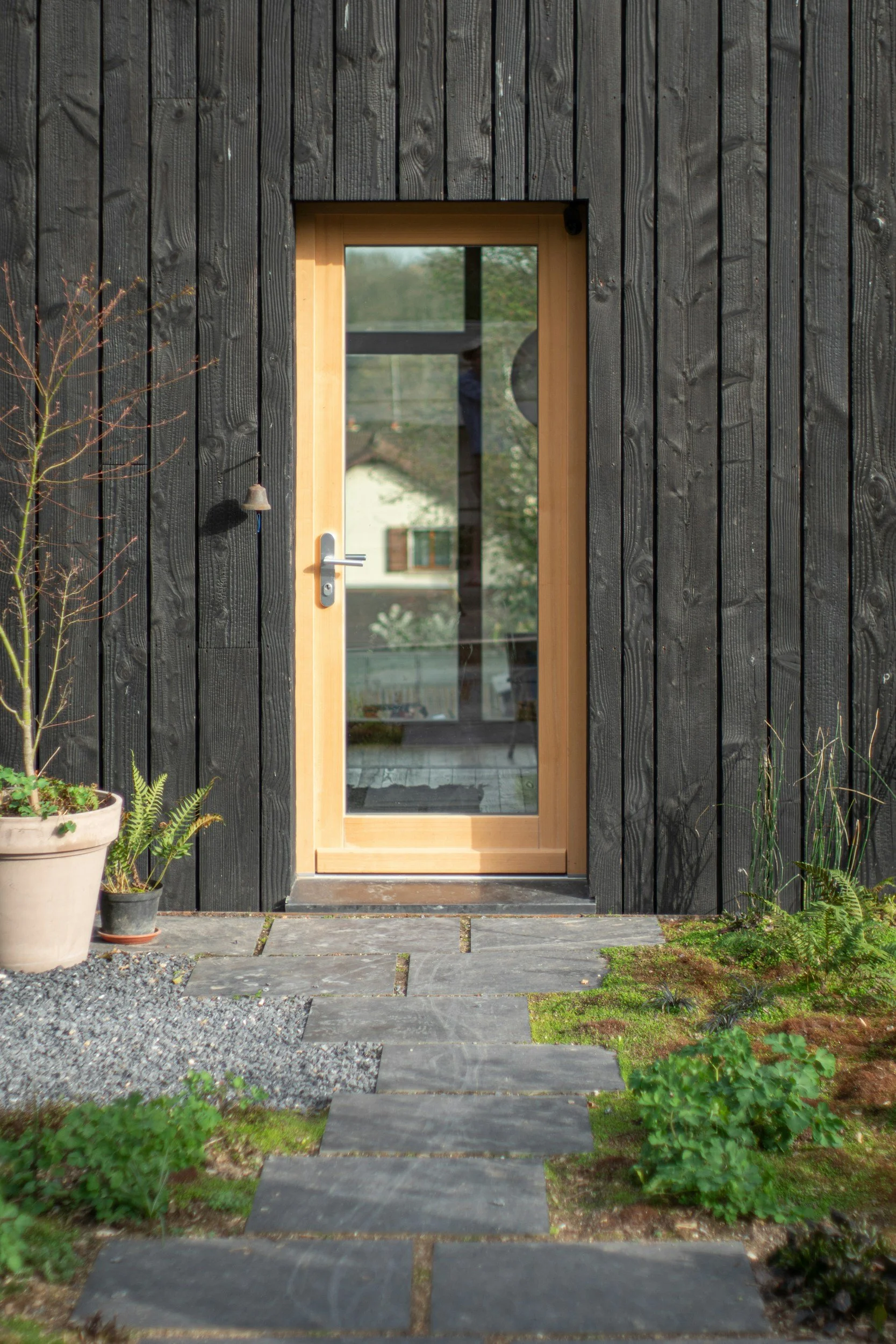 A black wooden house with a glass door framed in light wood, a small bell hanging to the left, and potted plants on the left side of the entrance. A stone pathway with patches of grass leads up to the door.