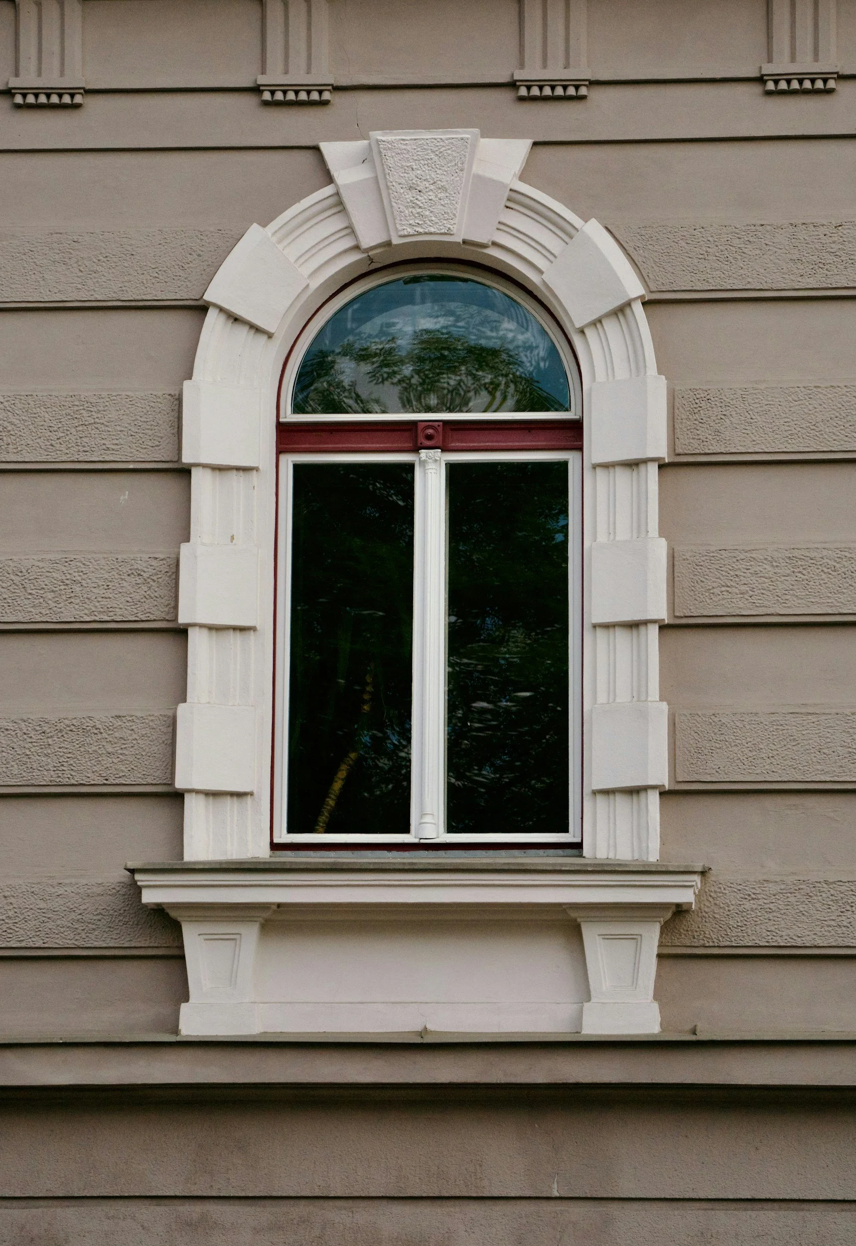 A rectangular window with an arched top on a beige building, decorated with white ornate trim and decorative molding.
