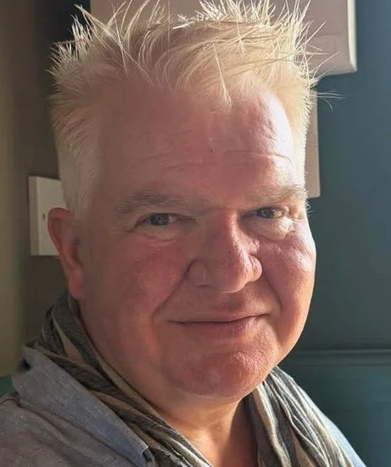 Close-up of a smiling middle-aged man with spiky blonde hair, wearing a gray shirt and a striped scarf, indoors near a blue wall.