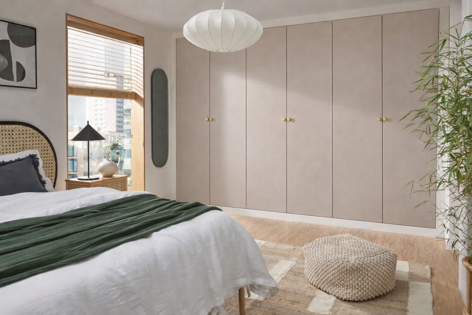 A bedroom with a bed, green blanket, black pillow, nightstand with black lamp, white vase, and plant, a beige wardrobe, a window with blinds, a white ceiling lamp, a mirror, and a textured beige pouf on a striped rug.
