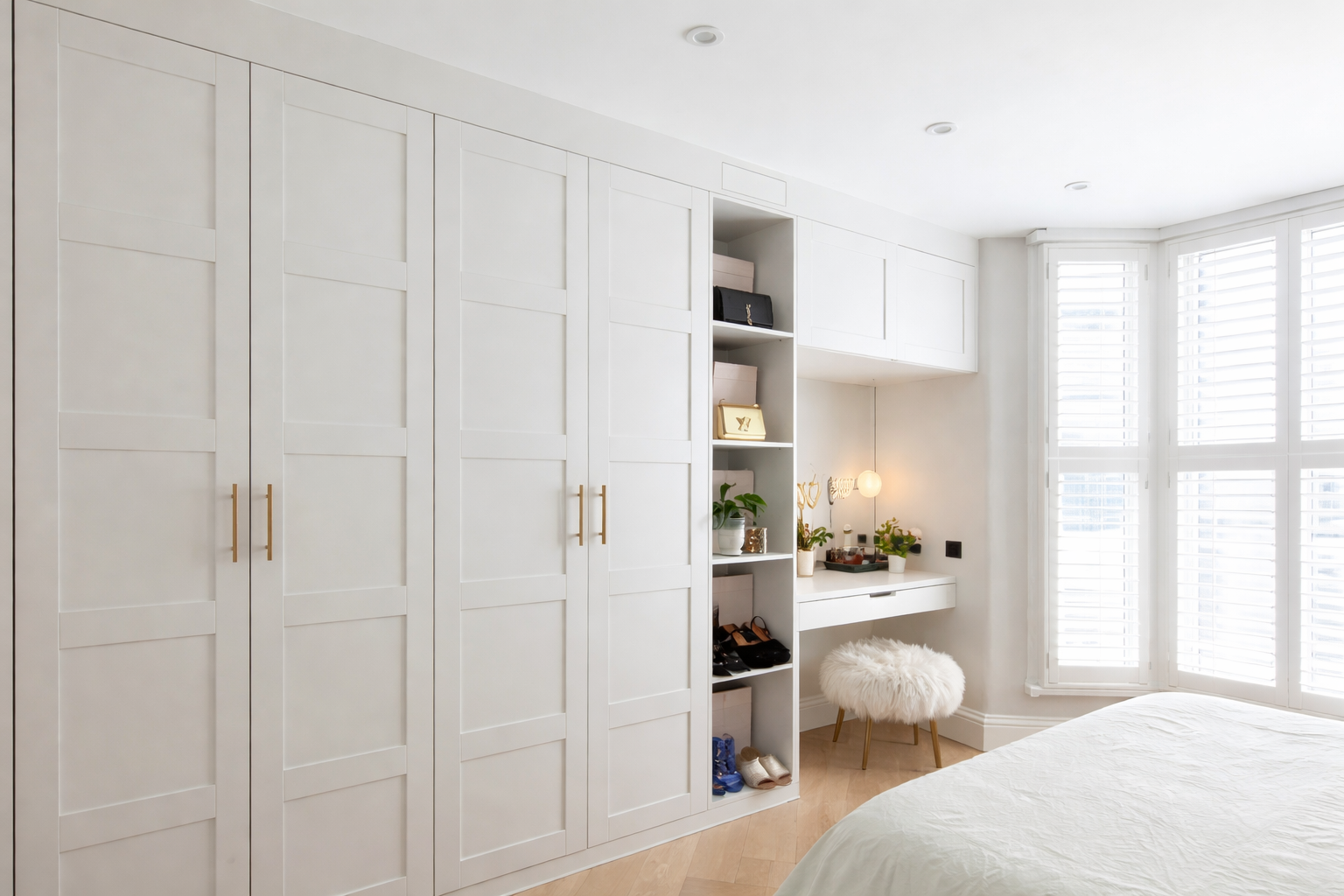 A bright bedroom with white built-in wardrobes, a small desk with plants and decorative items, a fluffy stool, and large windows with shutters.