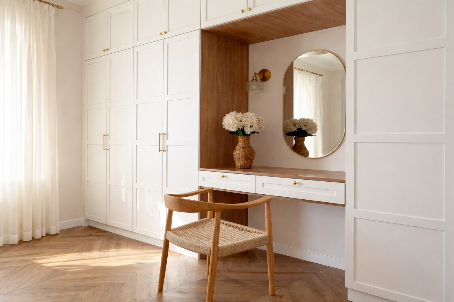 A cozy bedroom corner with built-in white wardrobes, a wooden vanity table with a mirror, a wicker vase filled with white flowers, and a wooden chair with woven seat. Light curtains and hardwood flooring complete the scene.