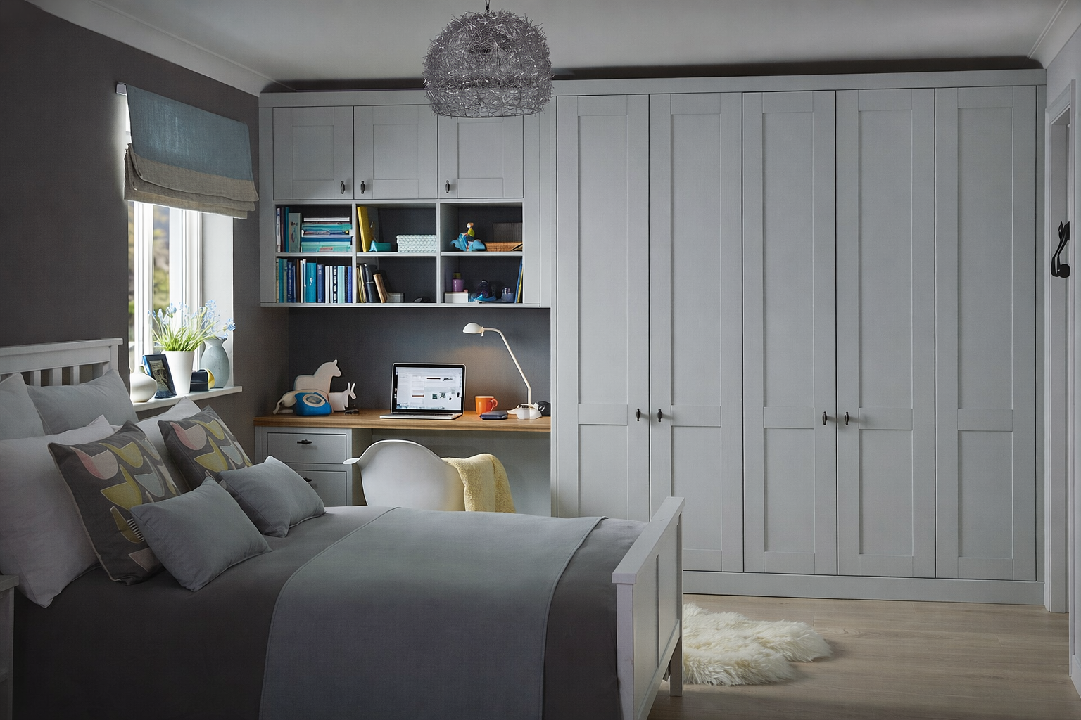 A bedroom with a white bed, decorative pillows, a desk with a laptop, books, and toys, a window with a blue window shade, a white chair with a yellow blanket, large white wardrobe, and a fluffy white rug, illuminated by natural light.