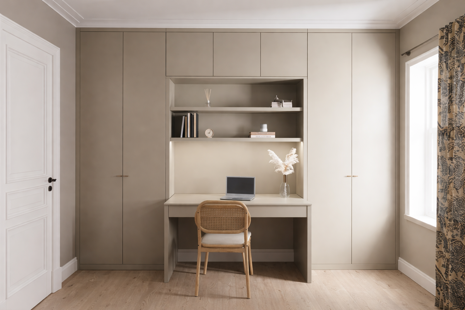 Home office nook with a built-in beige desk and cabinets, a laptop, a vase with pampas grass, and a wooden chair, in front of a window with patterned curtains.
