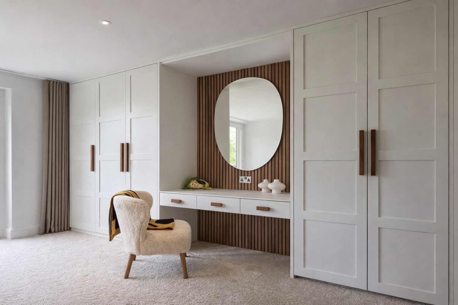 Modern bedroom with white and wood paneling, a large white wardrobe, an oval mirror, a white desk, a plush beige chair, and beige carpeting.