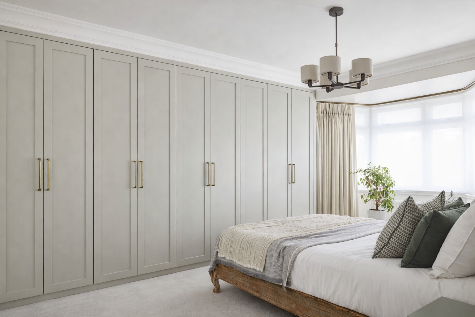 A bedroom with a large wardrobe, a bed with pillows and blankets, a window with curtains, a potted plant, and a ceiling chandelier.