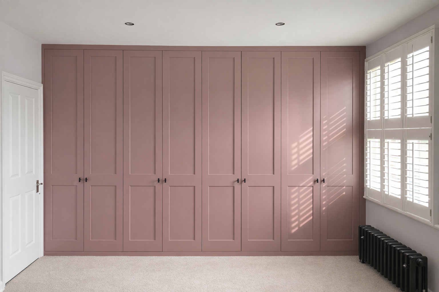 A room with a large pink wardrobe, white door on the left, window with white shutters on the right, and a black radiator below the window, with sunlight casting shadows through the shutters.