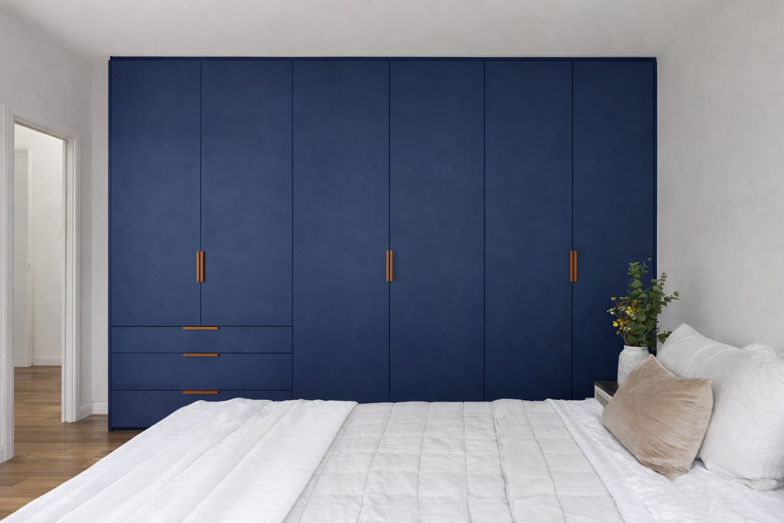 A bedroom with a large navy blue wardrobe, a white bed with pillows, and a nightstand with a vase of flowers.
