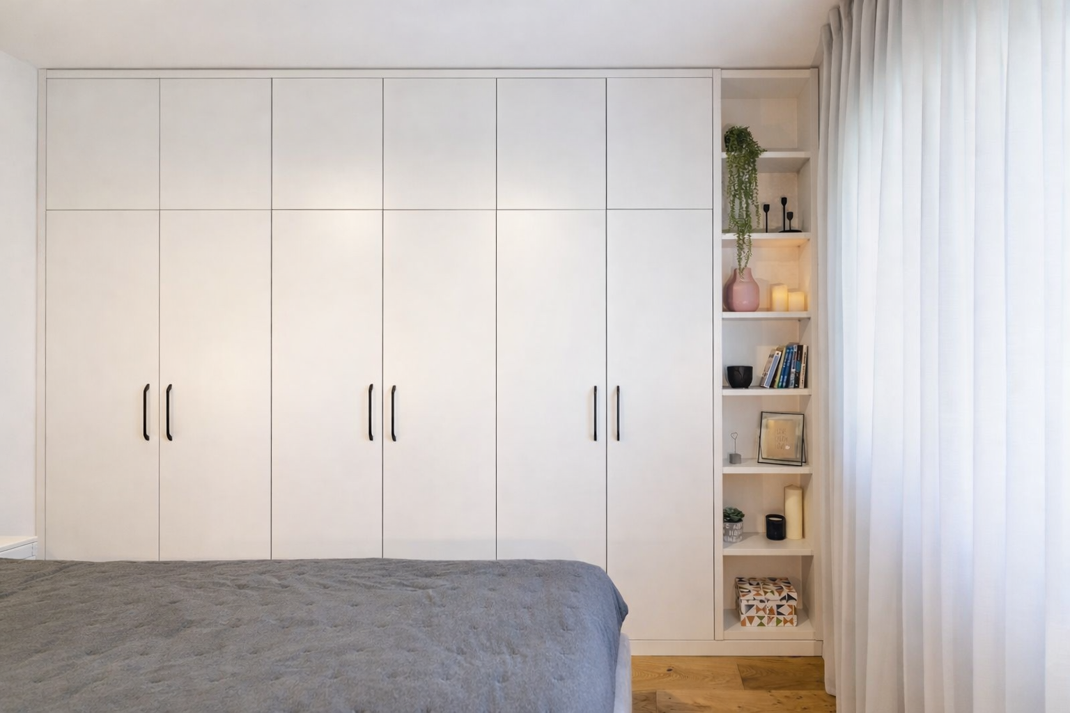 A bedroom with a large white built-in wardrobe, a gray bedspread, and an open white bookshelf with decorative items, books, and candles, next to white curtains letting in natural light.
